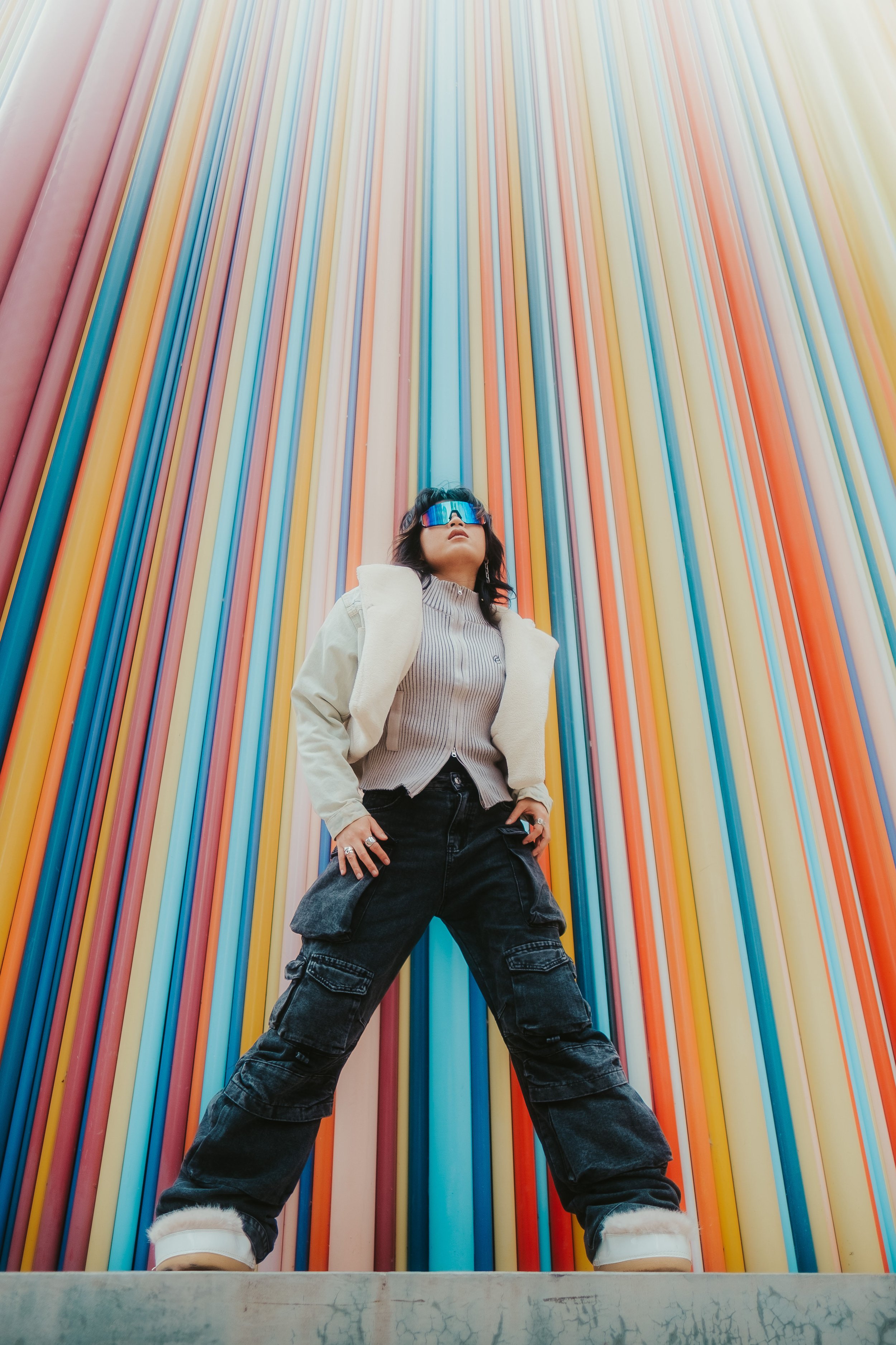 Mulher com roupas casuais, jaqueta e calças largas, usando óculos de proteção visual, posando em frente a uma parede de tubos coloridos Verticalmente.