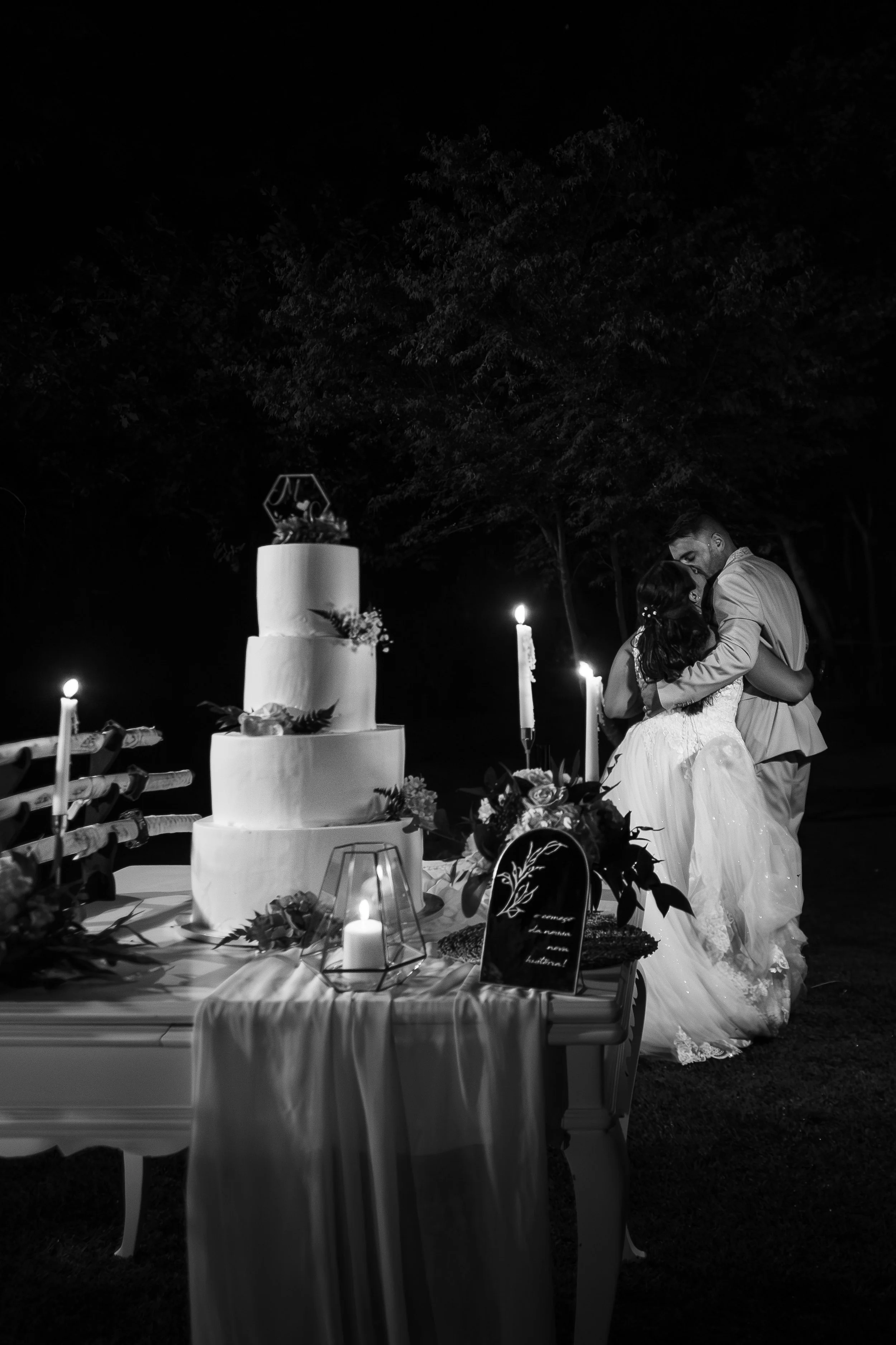 Casal de casamento dançando ao lado de um bolo de casamento de quatro andares, decorado com flores, em duas velas acesas, em uma cerimônia ao ar livre à noite.