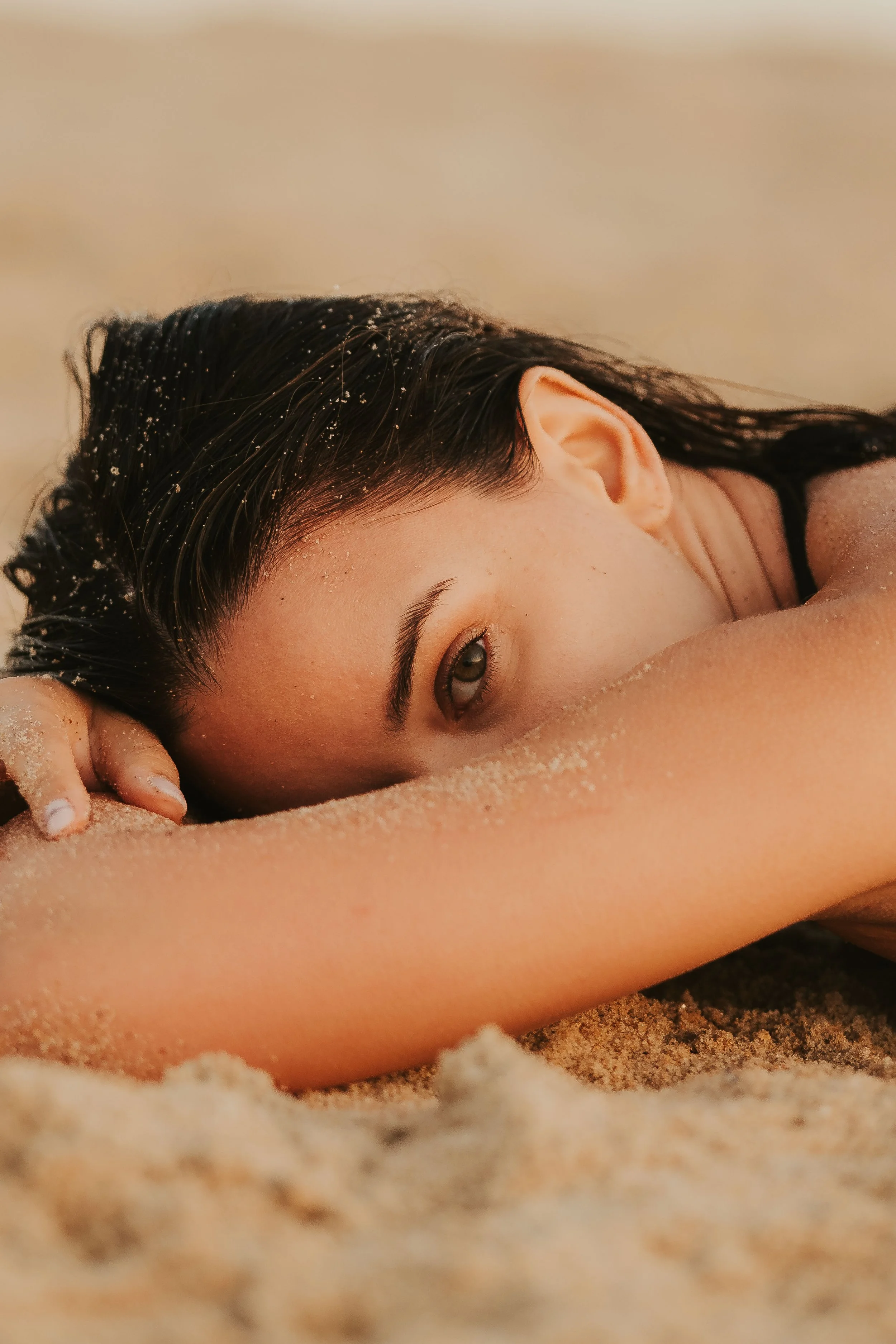 Mulher deitada na areia em um ambiente de praia, com cabelo escuro e olhos claros, parcialmente coberta de areia.