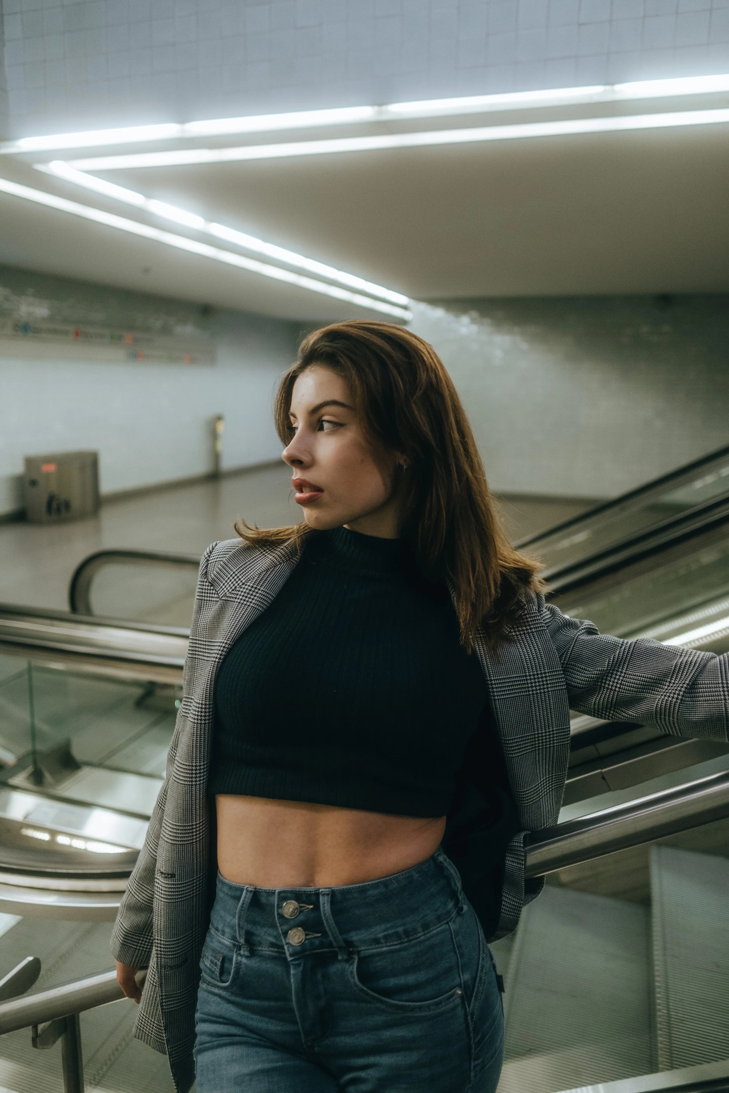 Mulher adolescente com cabelo castanho, usando blusa preta de manga longa, jaqueta de tweed cinza e jeans, posando ao lado de uma escada rolante em uma estação de metrô ou metrô subterrâneo