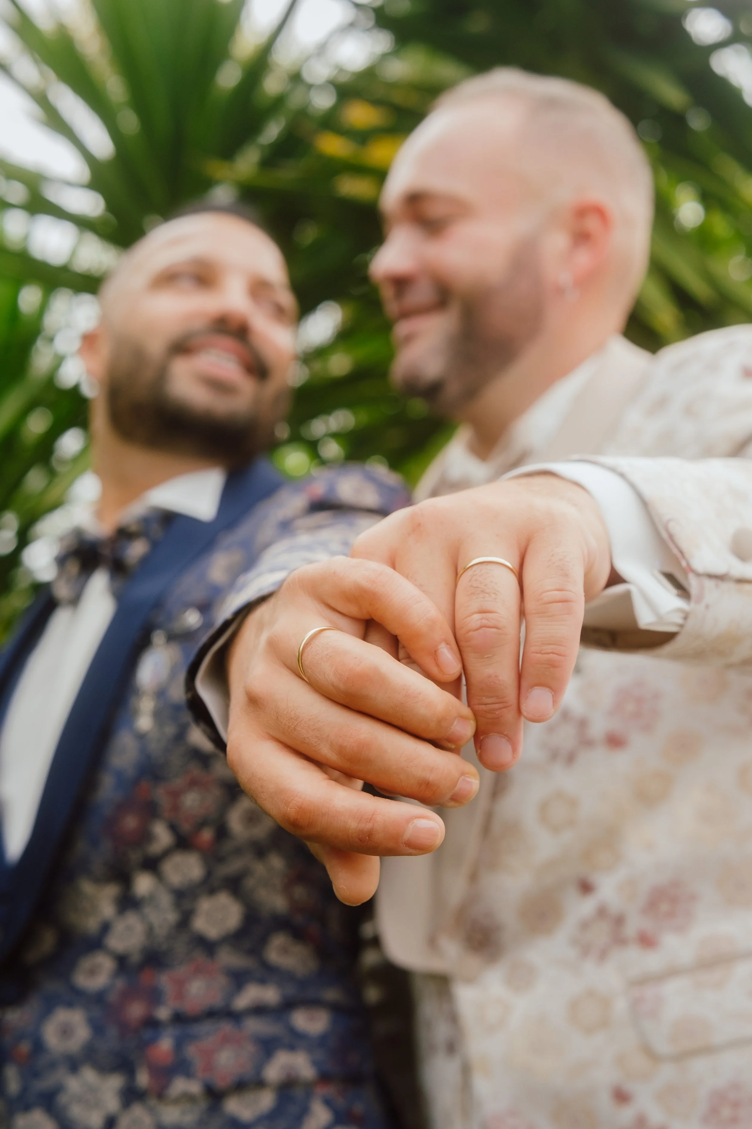 Homens gays segurando as mãos, usando alianças de casamento, sorrindo um para o outro, com fundo de plantas verdes.