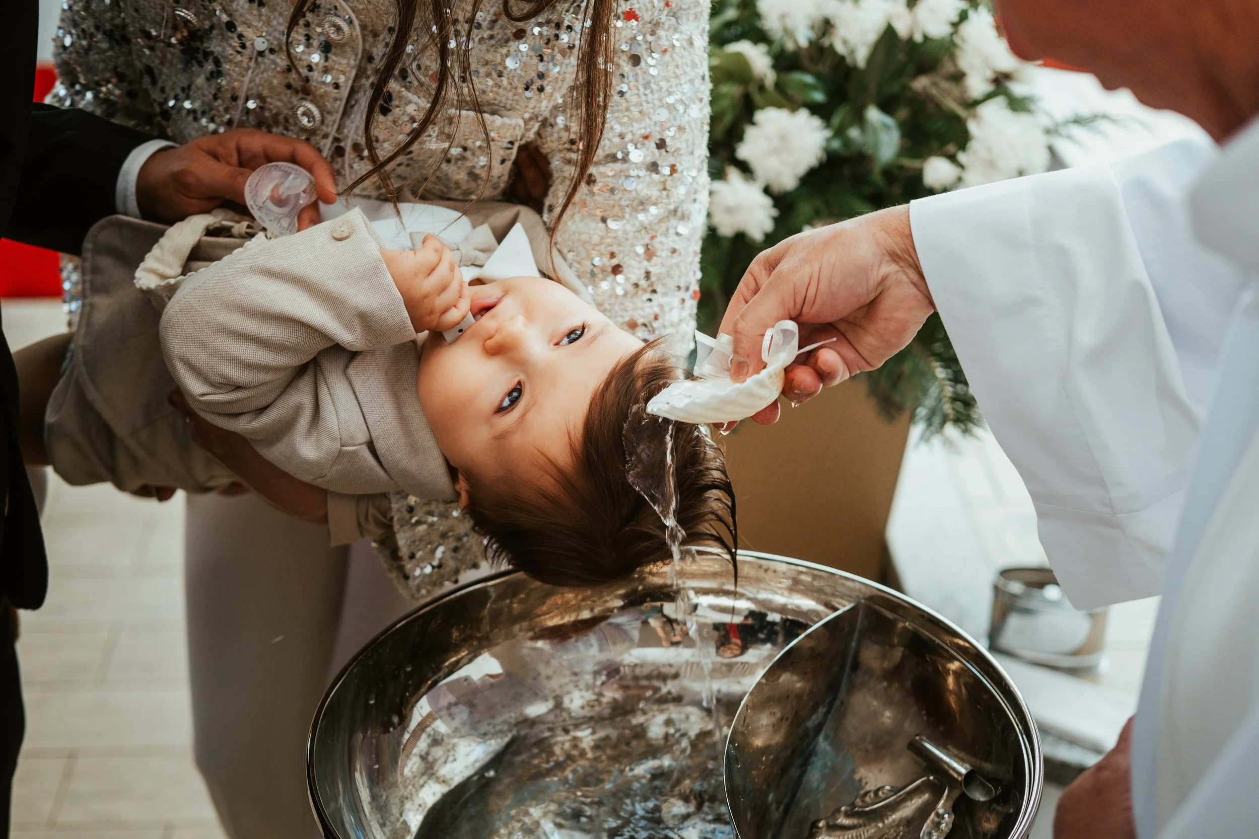 Bebê sendo batizado com água na testa por um padre, com os pais segurando o bebê. Fundo com flores brancas e árvores verdes.