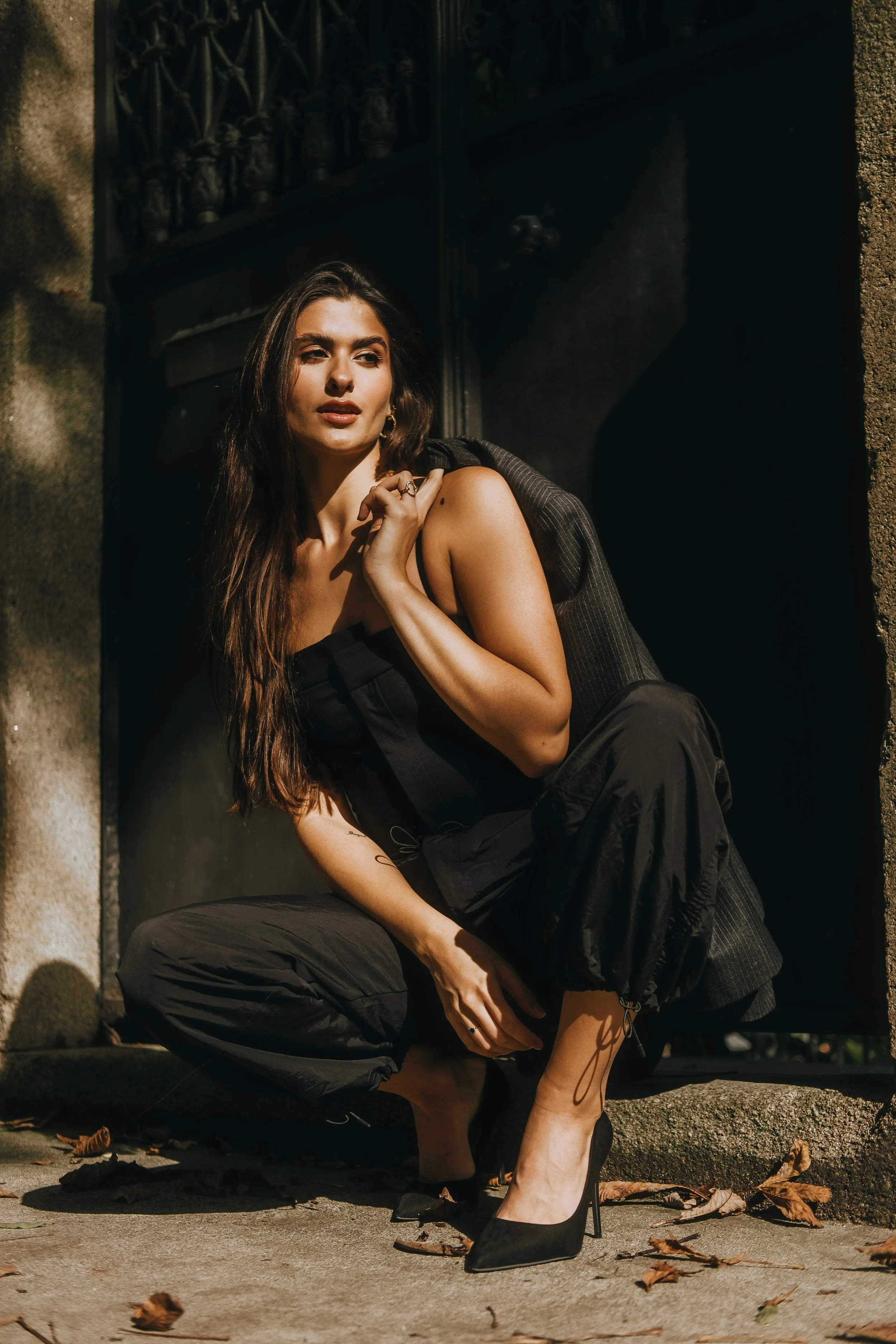 Mulher com roupas pretas, segurando um saco nos ombros, agachada ao lado de uma rua com folhas secas no chão, sob luz solar