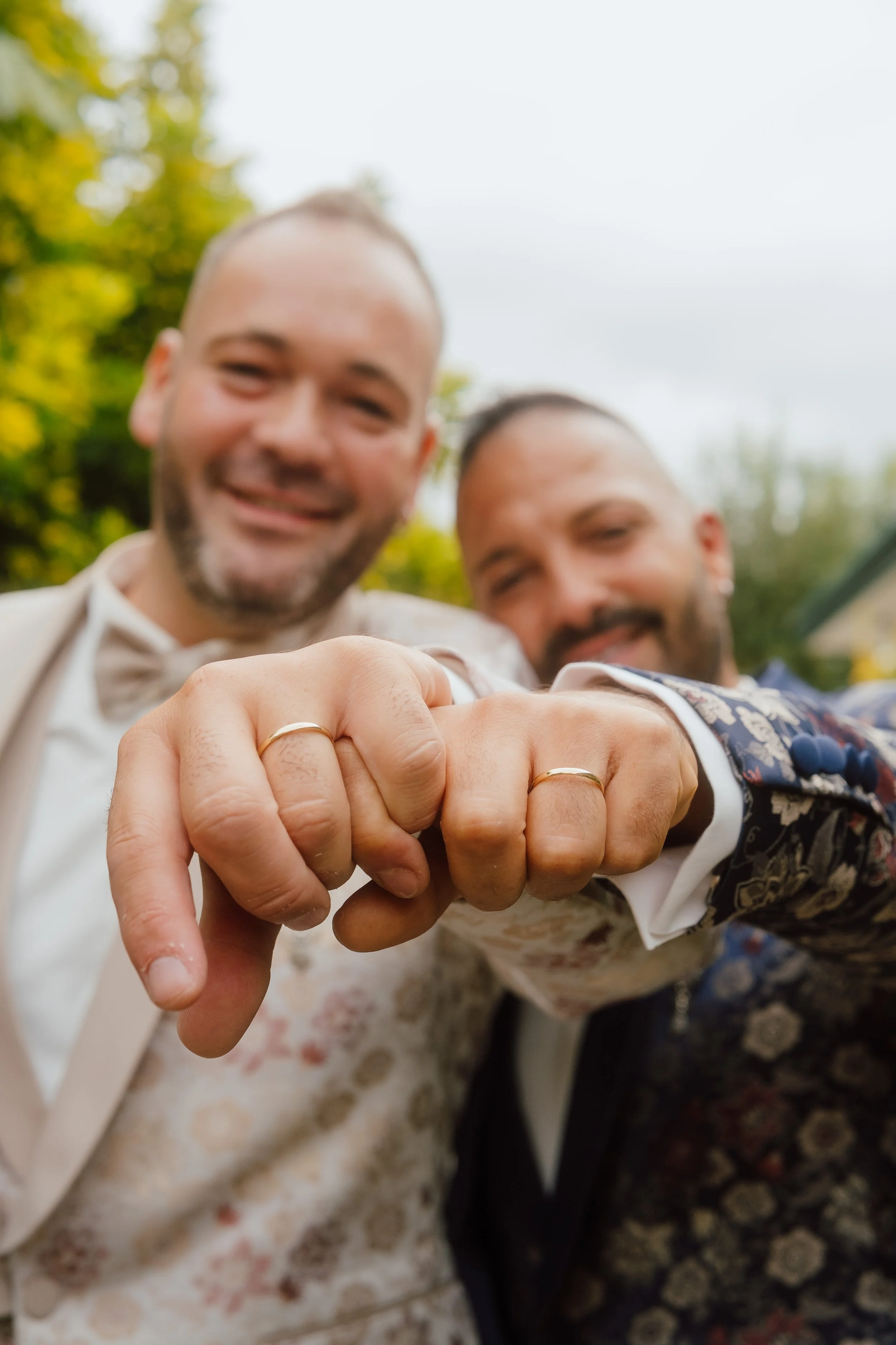 Casais de même gênero exibindo alianças de casamento em um abraço, sorrindo e com fundo de árvores verdes.
