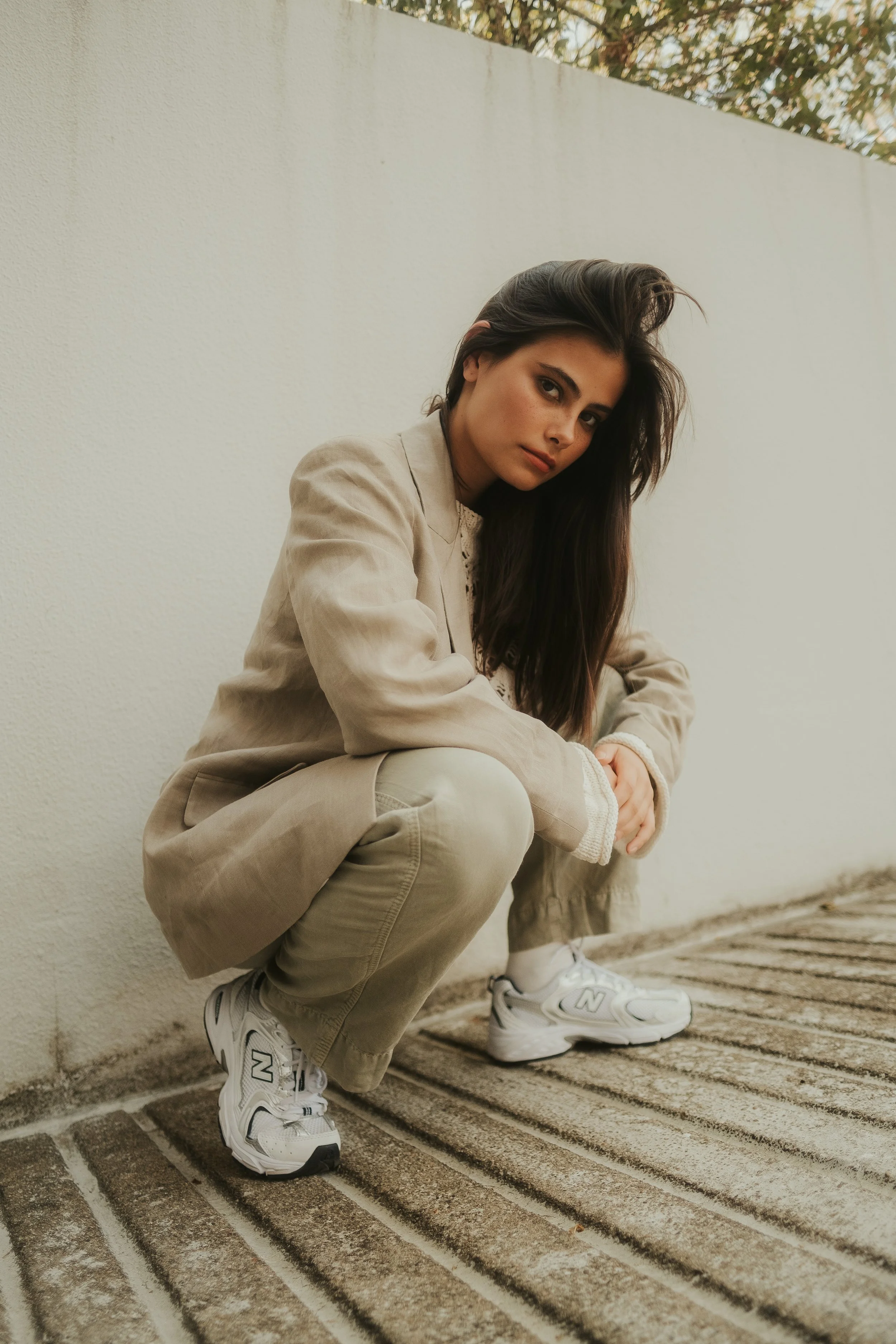 Rapaz de cabelo castanho escuro, usando blazer bege, calça bege, tênis branco e camisa de manga longa, crouching next to a white wall, outdoors.