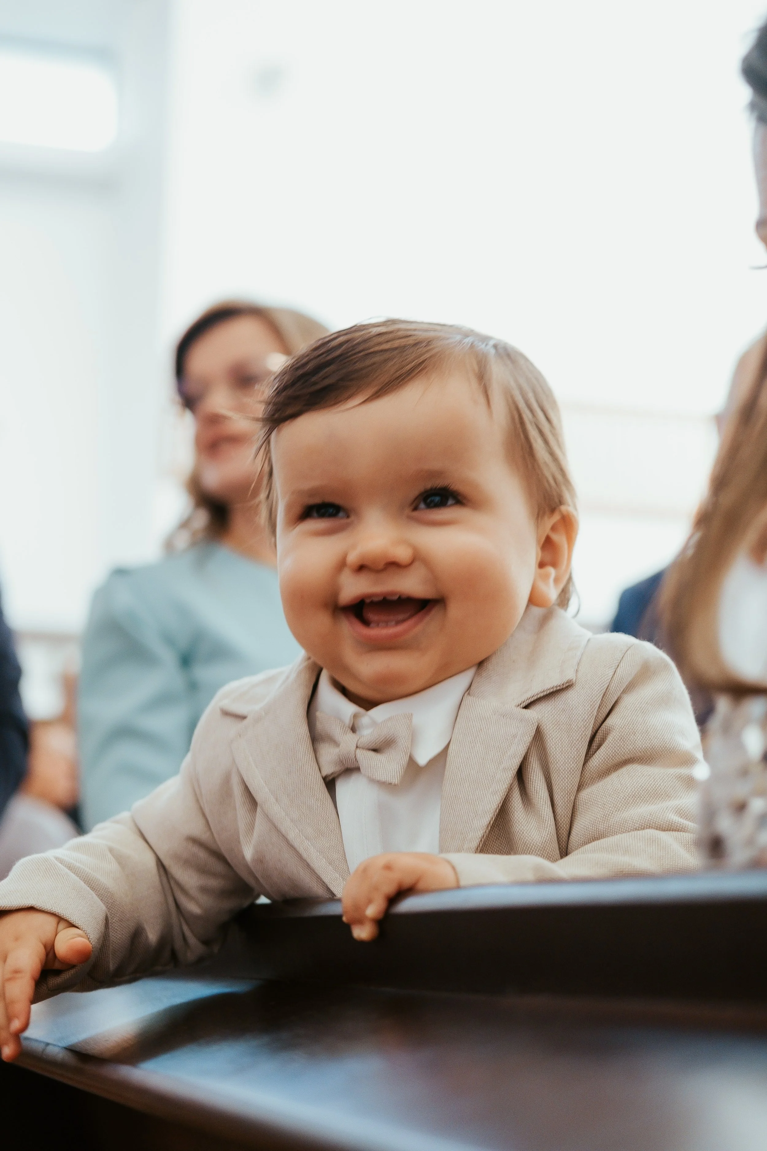 Menino sorridente com terno e gravata borboleta, em um ambiente iluminado, sendo observado por adultos ao fundo.