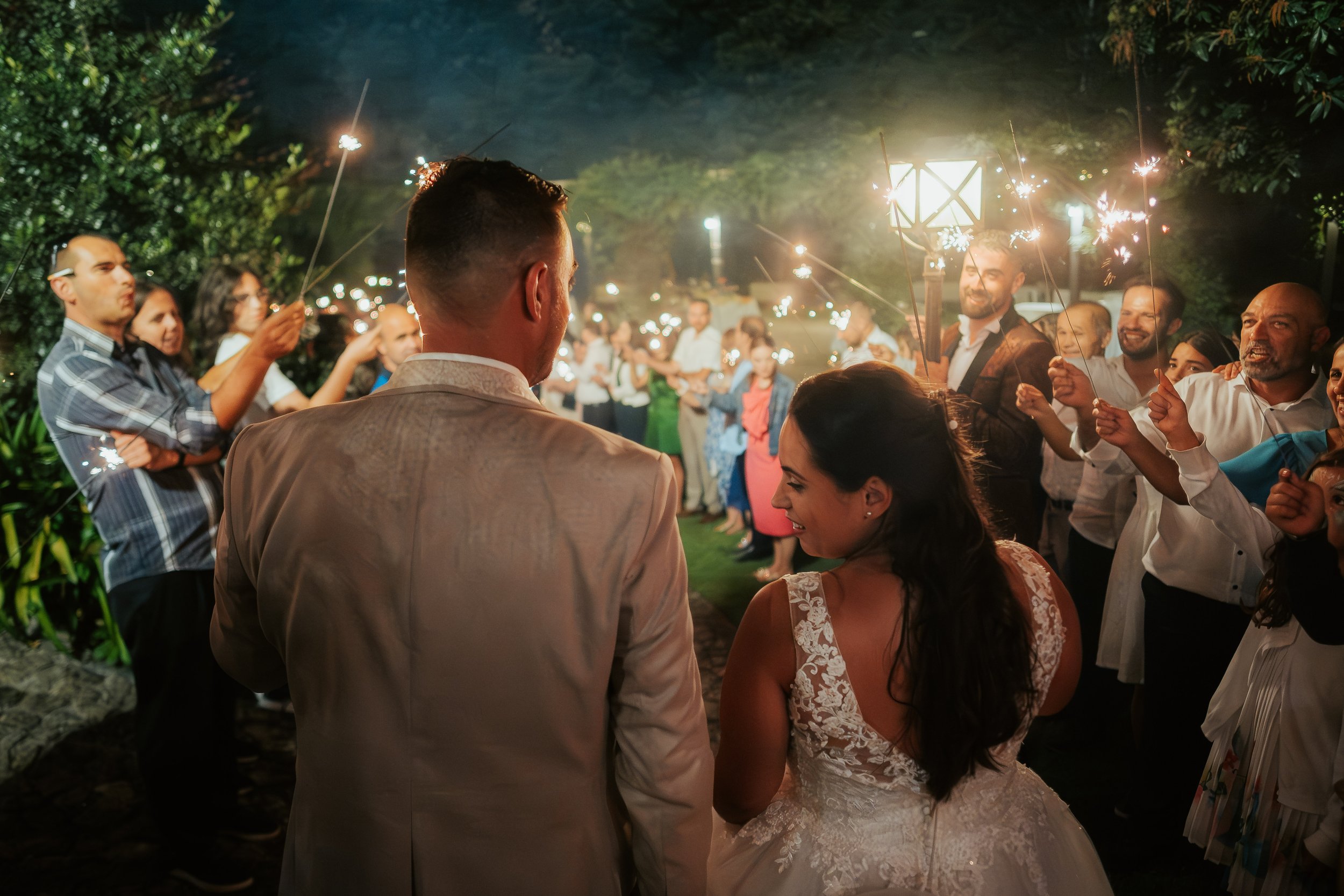 Casal de noivos com convidados em uma festa de casamento ao ar livre, segurando tochas com fogos de artifício, à noite.