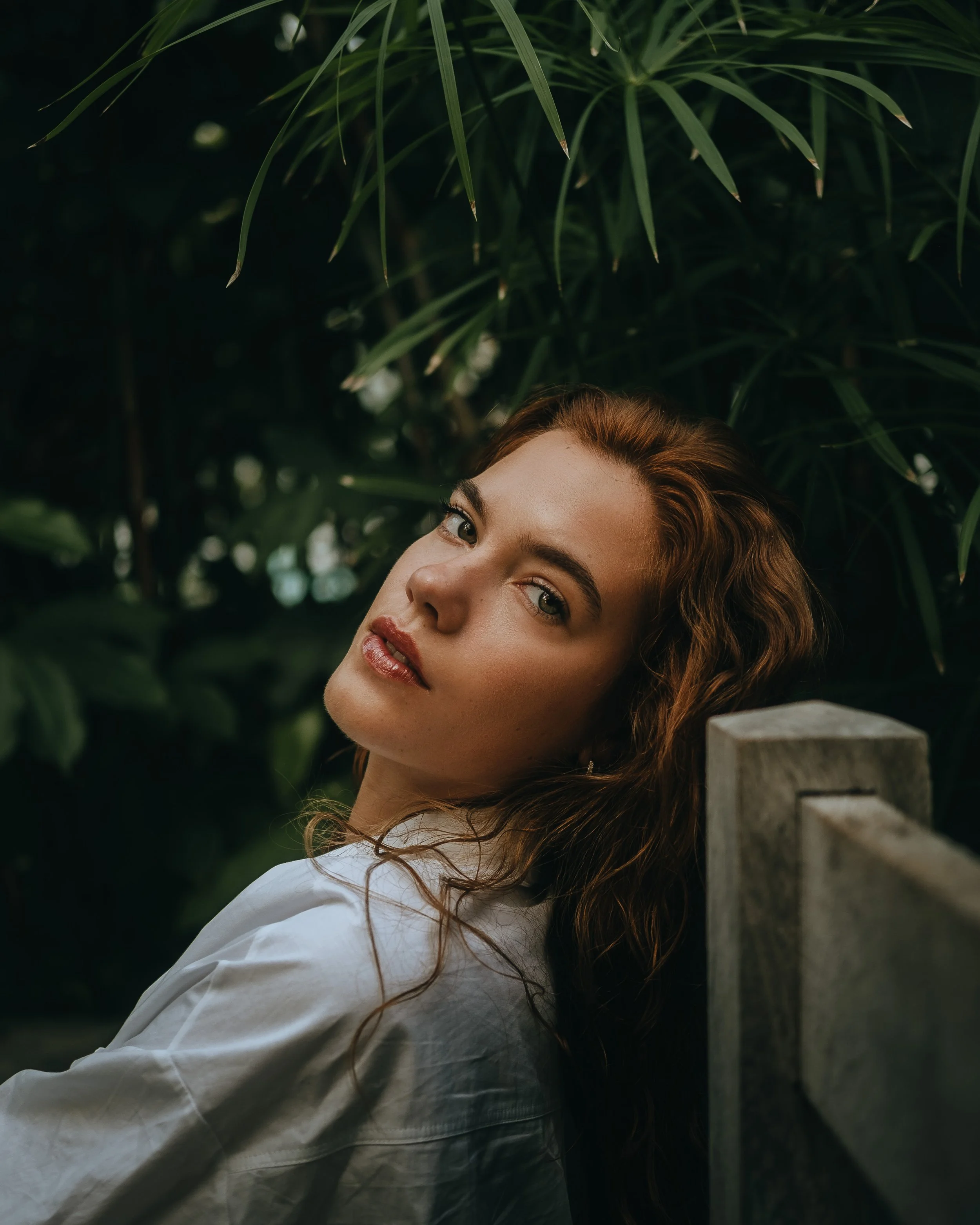 Retrato de uma mulher de cabelos castanhos encarando a câmera, de cabeça levemente inclinada, com fundo de folhas verdes.