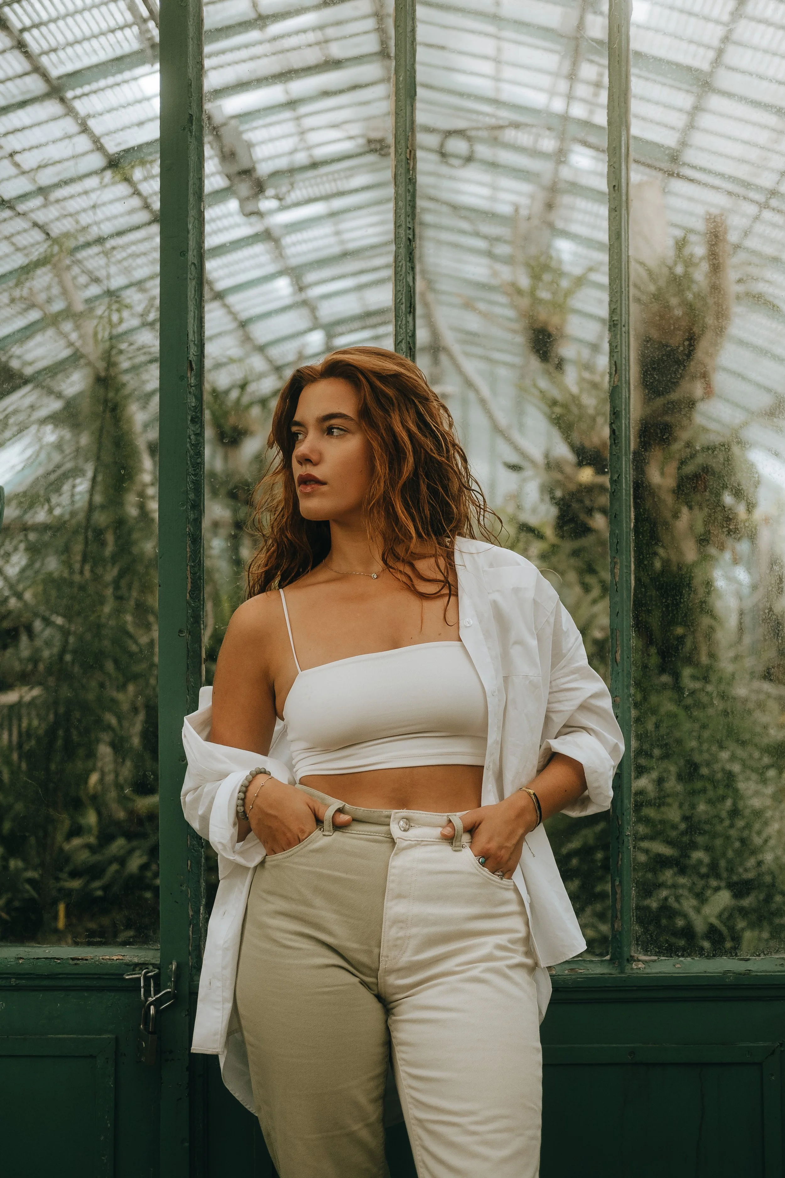 Mulher de cabelos castanhos ondulados, usando blusa branca de alça fina e calça bege, com uma camisa branca meio aberta, posando em frente a uma estufa de vidro com plantas ao fundo.