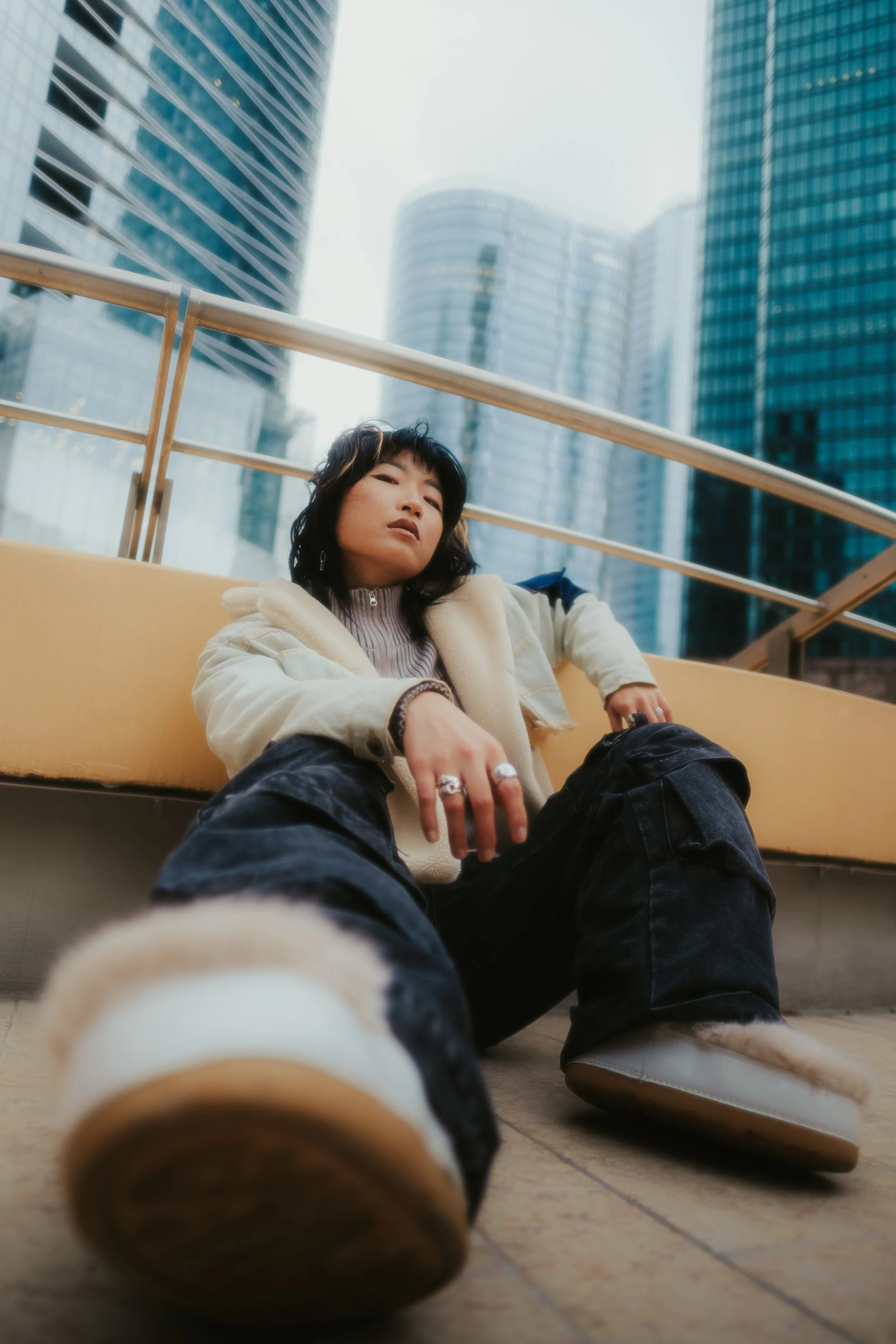 Jovem de cabelo escuro sentada no chão de um espaço urbano com prédios modernos ao fundo, usando roupas casuais e botas com forro de pelúcia, olhando para a câmera com expressão de confiança.