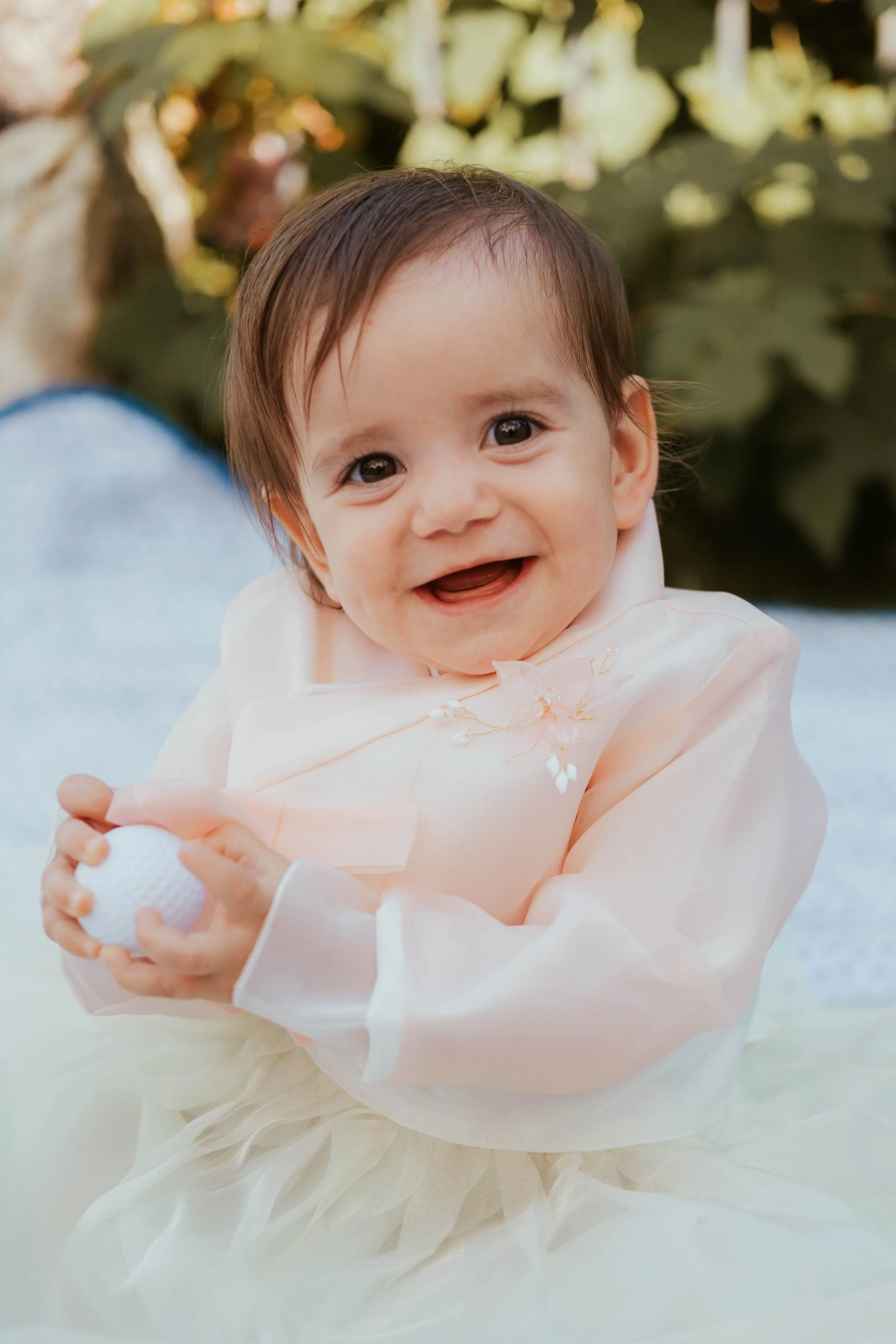 Bebê sorridente de vestido claro segurando uma bola de golfe, ao ar livre, com folhagem ao fundo.