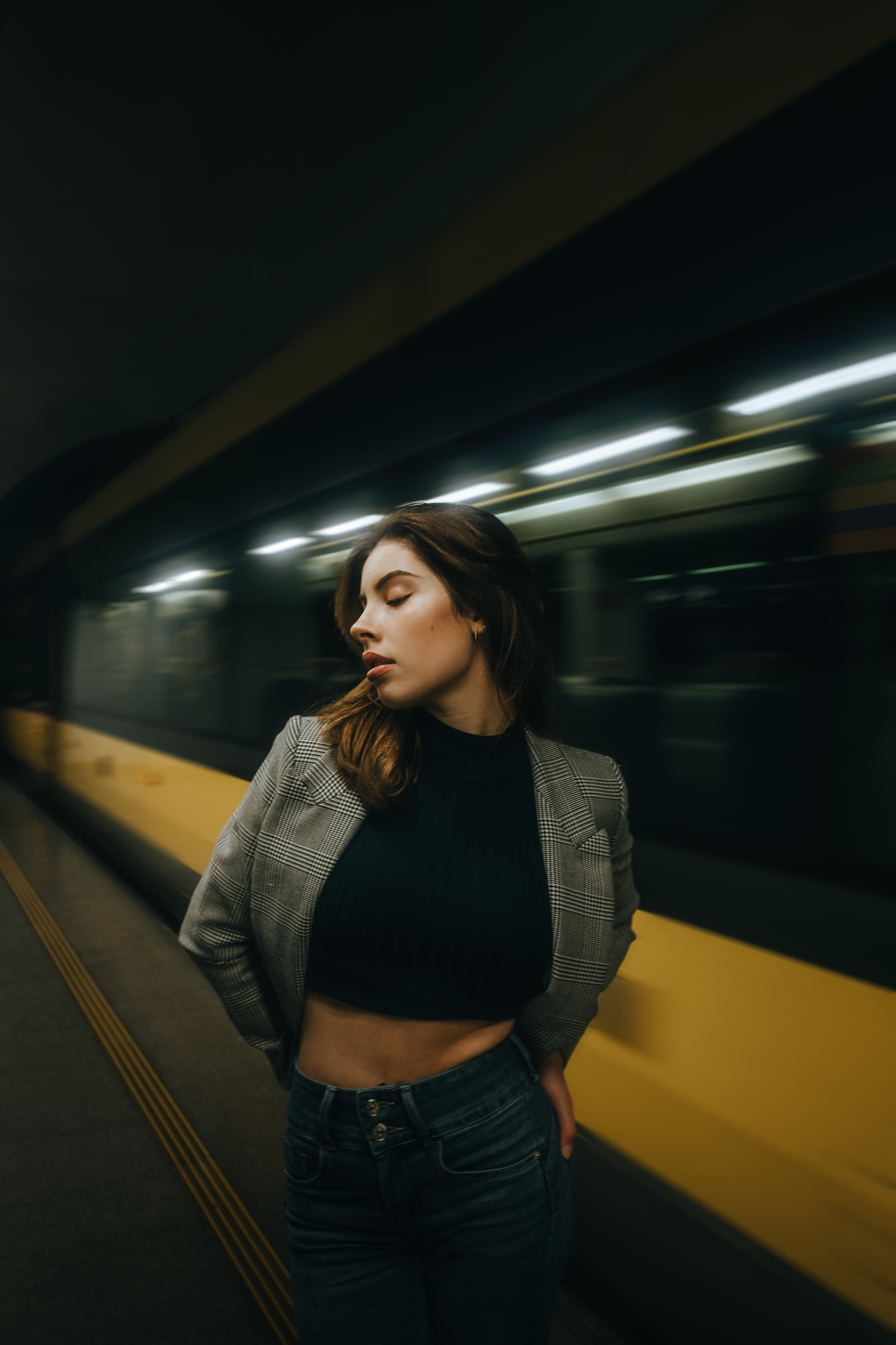 Mulher com blusa preta e blazer xadrez em estação de metrô, posando com olhos fechados, na plataforma do metrô em movimento ao fundo.