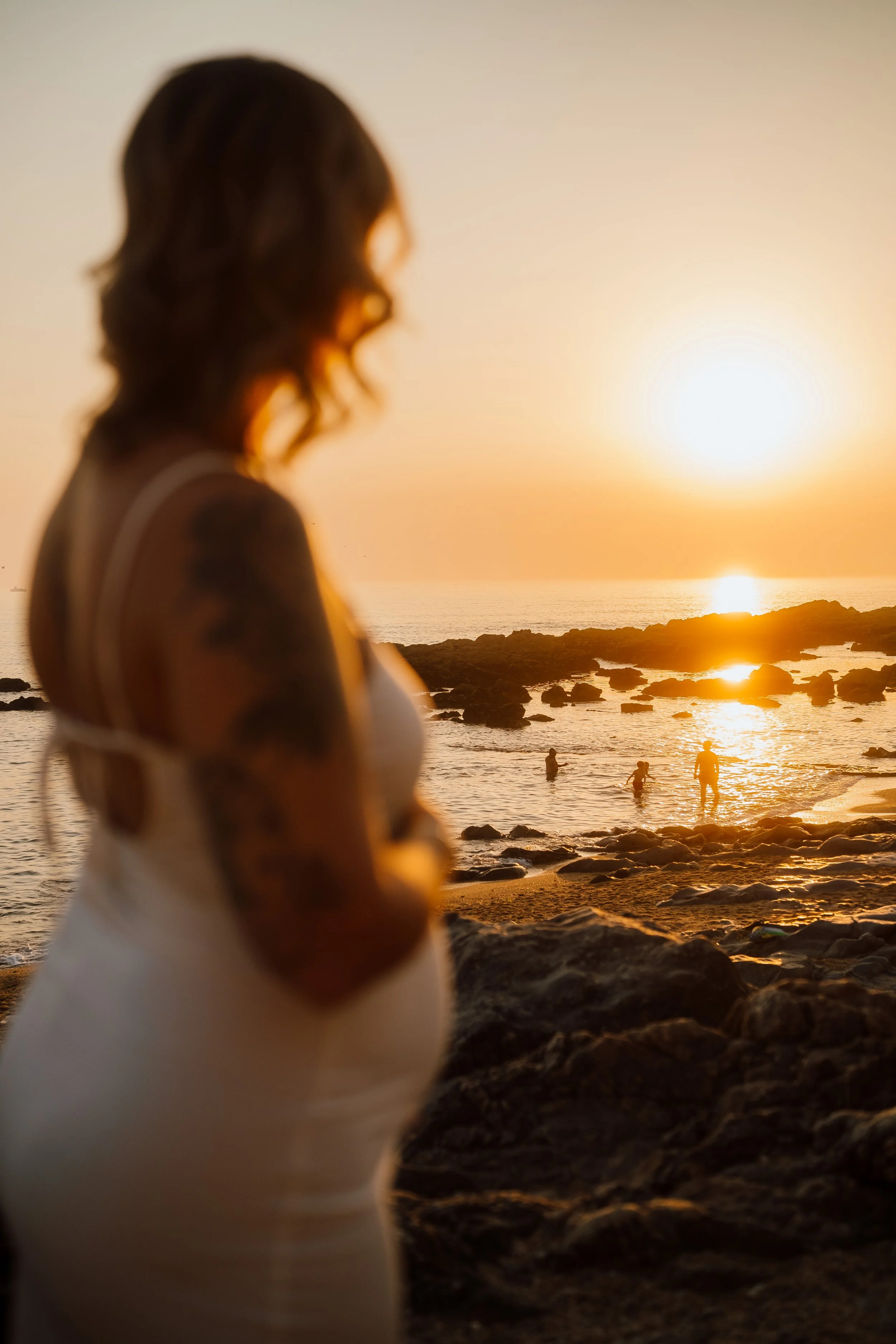 Mulher com tatuagem na braço de cabelo castanho, vestindo roupa branca, de costas, na praia ao pôr do sol, com pessoas ao fundo na água.