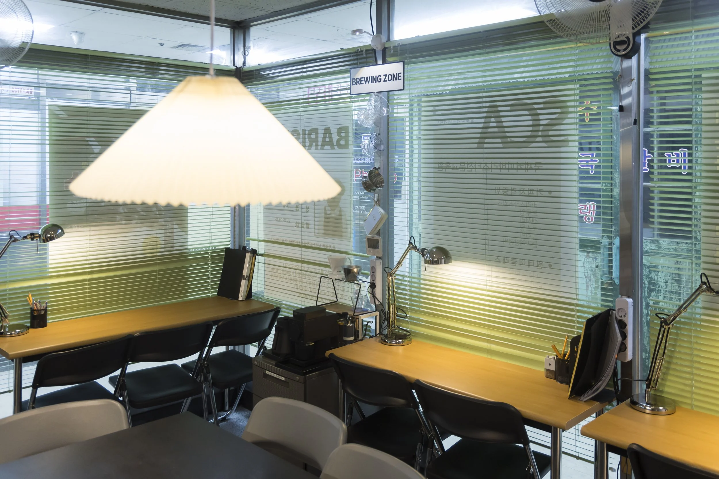 Interior view of a cafe with green blinds on large windows, a hanging lamp, desks with desk lamps, notebooks, pens, and electronic devices, and a sign indicating a brewing zone.
