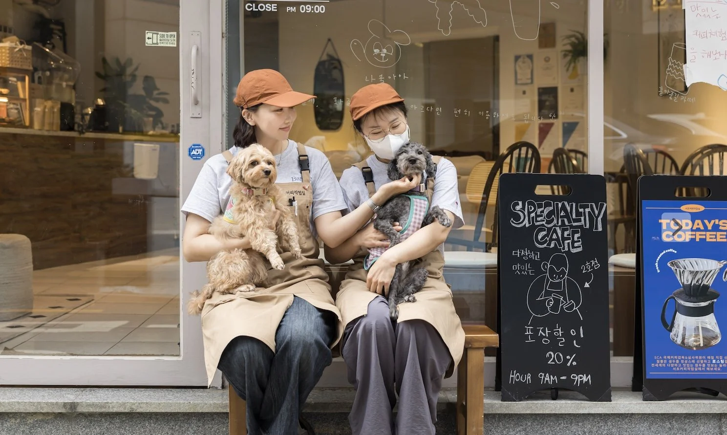 Two women sitting outside a cafe, each holding a small dog. One woman is wearing glasses and a face mask, and the other is not. Both women are wearing beige aprons and orange hats. There are signs advertising the cafe's specials and coffee outside.