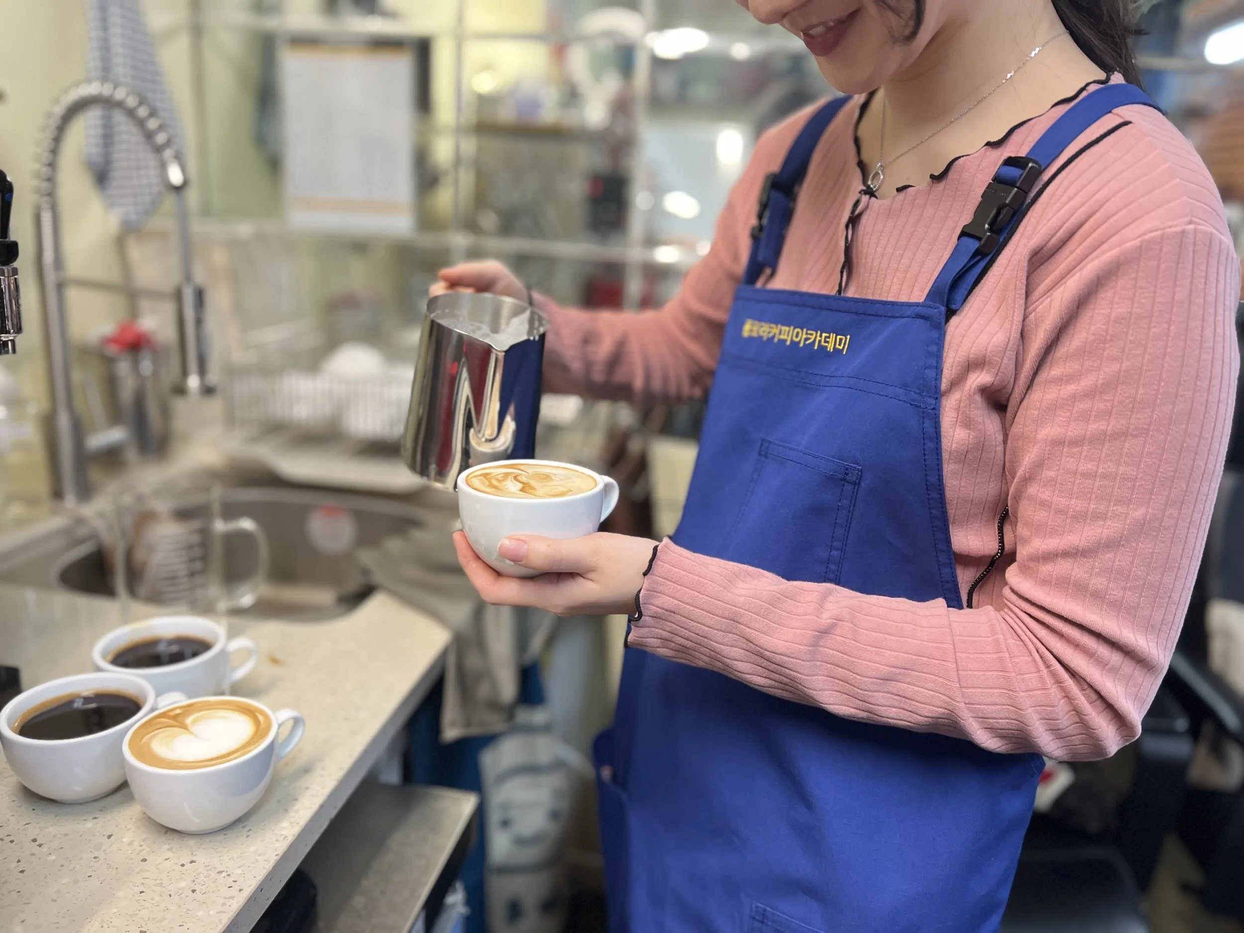 Barista pouring steamed milk into a cup of coffee with latte art. Three cups of coffee on the counter, with one having latte art, in a café setting.