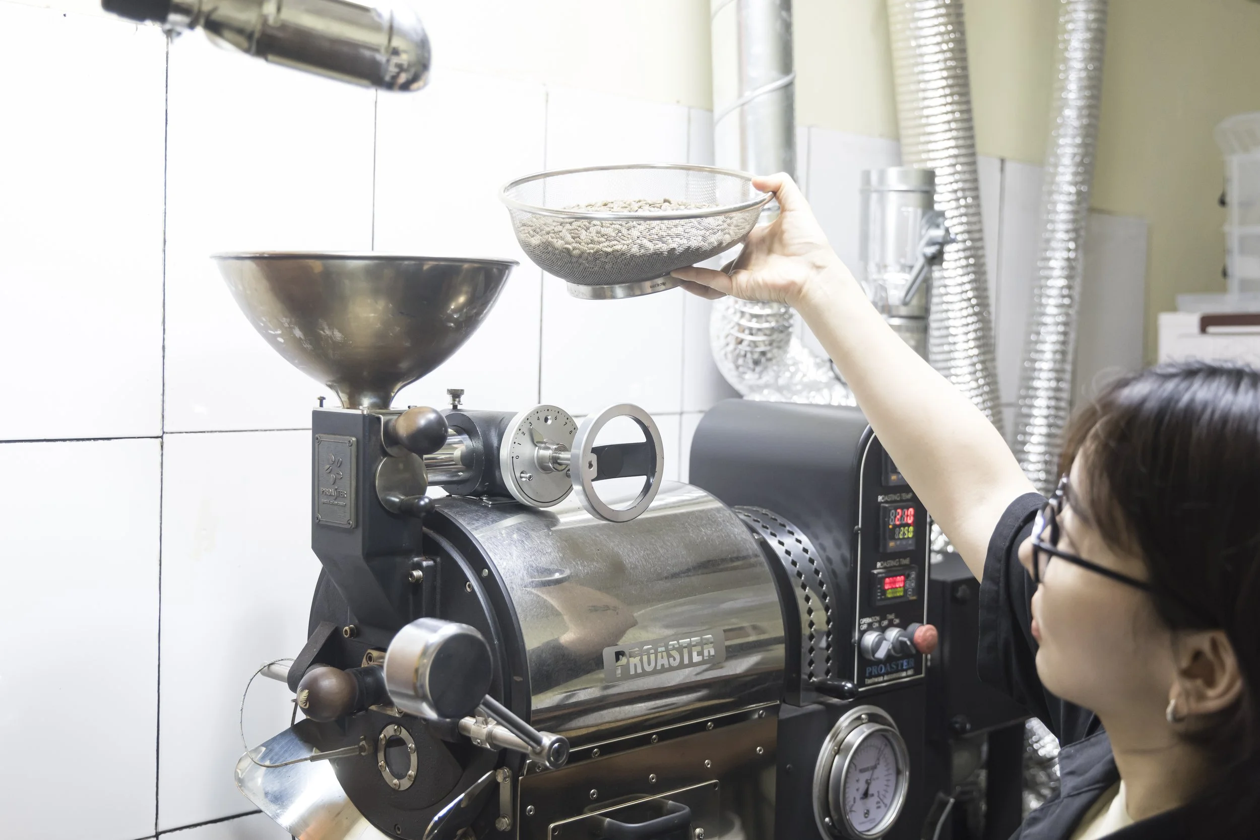 A person operating an industrial coffee roaster machine in a kitchen or coffee shop, placing a tray of green coffee beans into the machine.