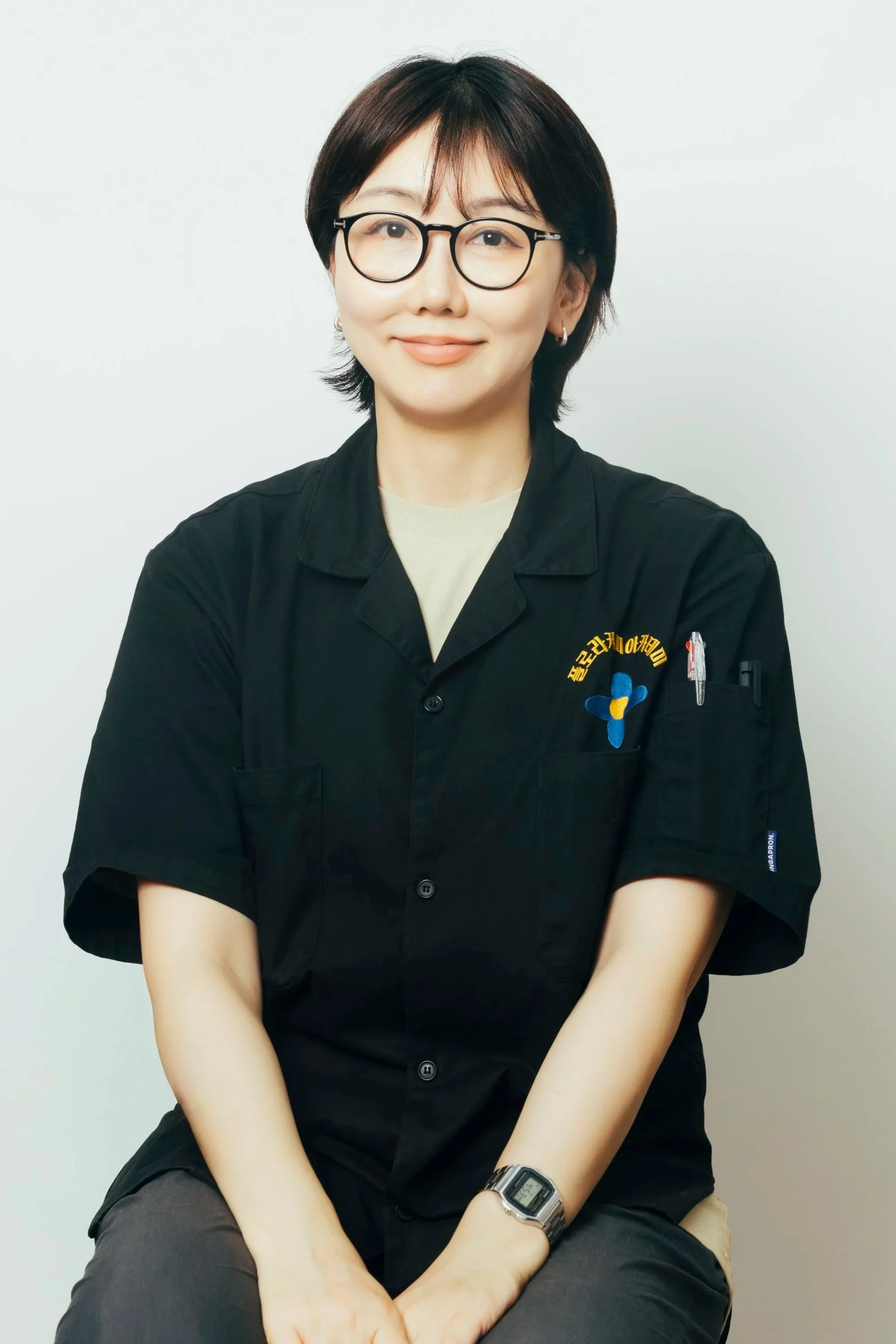 A woman with short dark hair, wearing glasses and a black uniform with a logo, sitting against a plain background.