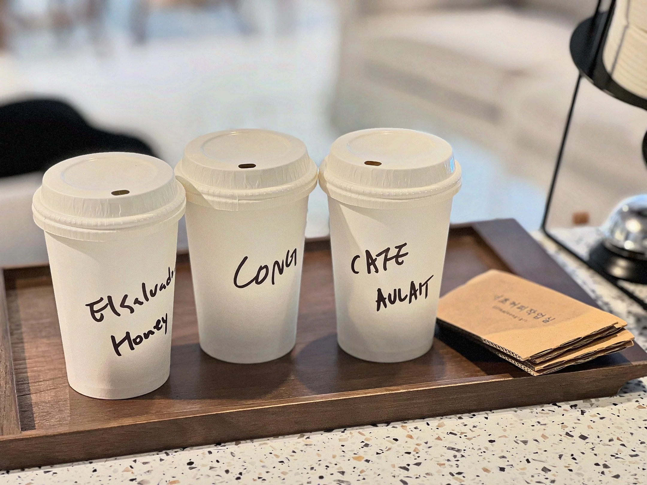Three to-go coffee cups with handwritten labels that read 'Elicited Honey,' 'CONG,' and 'CAFE AULAIT' placed on a wooden tray on a speckled countertop, with a brown paper bag nearby.