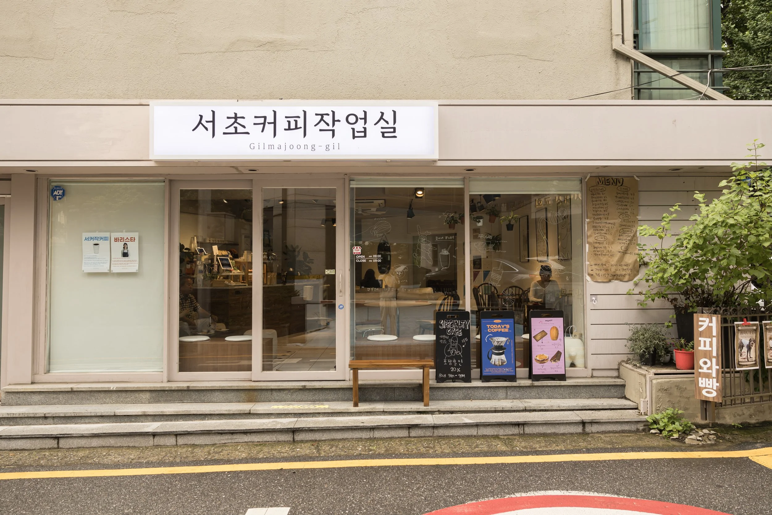 Front view of a coffee shop with a sign in Korean, glass windows, and outdoor signage advertising drinks and baked goods. Small wooden bench outside stairs with potted plants on the right.