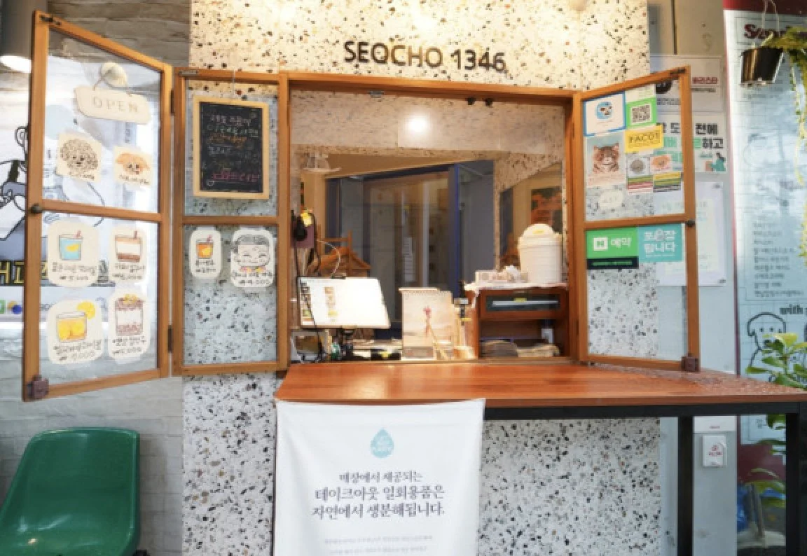 A small order counter at a cafe or restaurant with a wooden frame, featuring charts and signs, items for sale, and a computer or register, with a sign indicating contactless payment options.