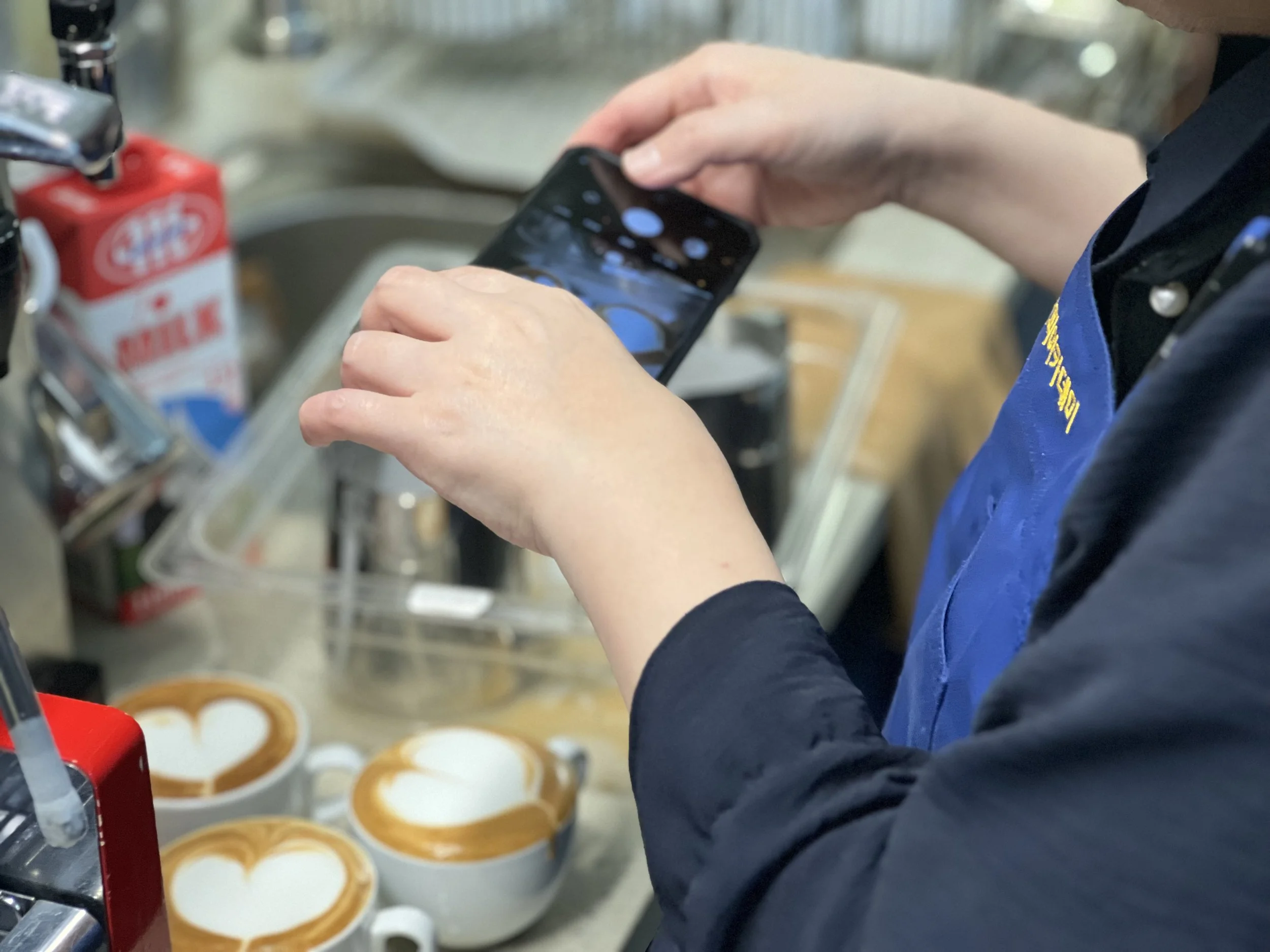 Barista taking a photo of three cappuccinos with heart-shaped latte art using a smartphone in a coffee shop.