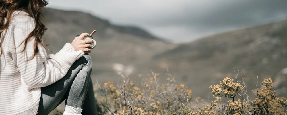 Woman in a white sweater sitting outdoors with a mug, overlooking a mountain landscape