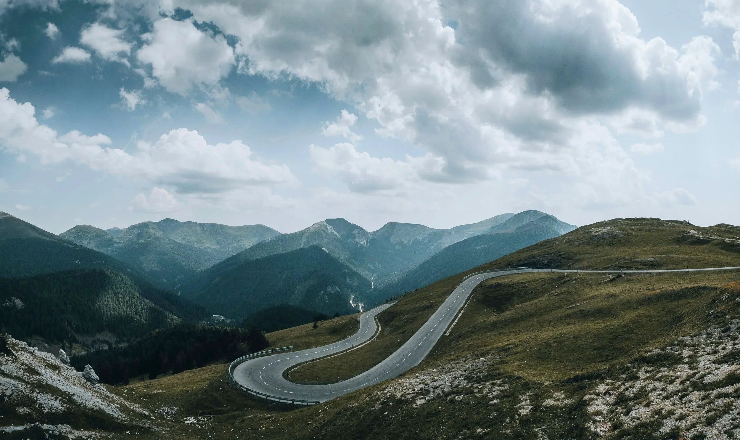 Winding mountain road through alpine terrain with forested valleys, rugged peaks, and dramatic clouds