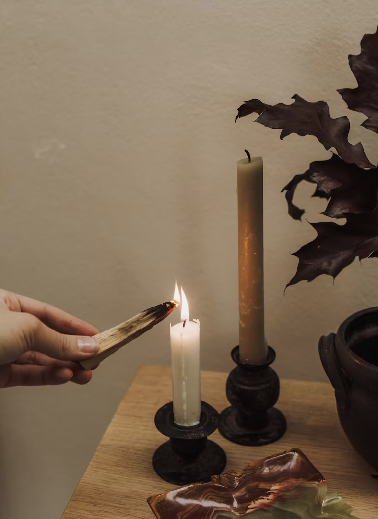 Hand lighting a white taper candle beside a tall brown candle, dark candlesticks, and dried leaves on a wooden table