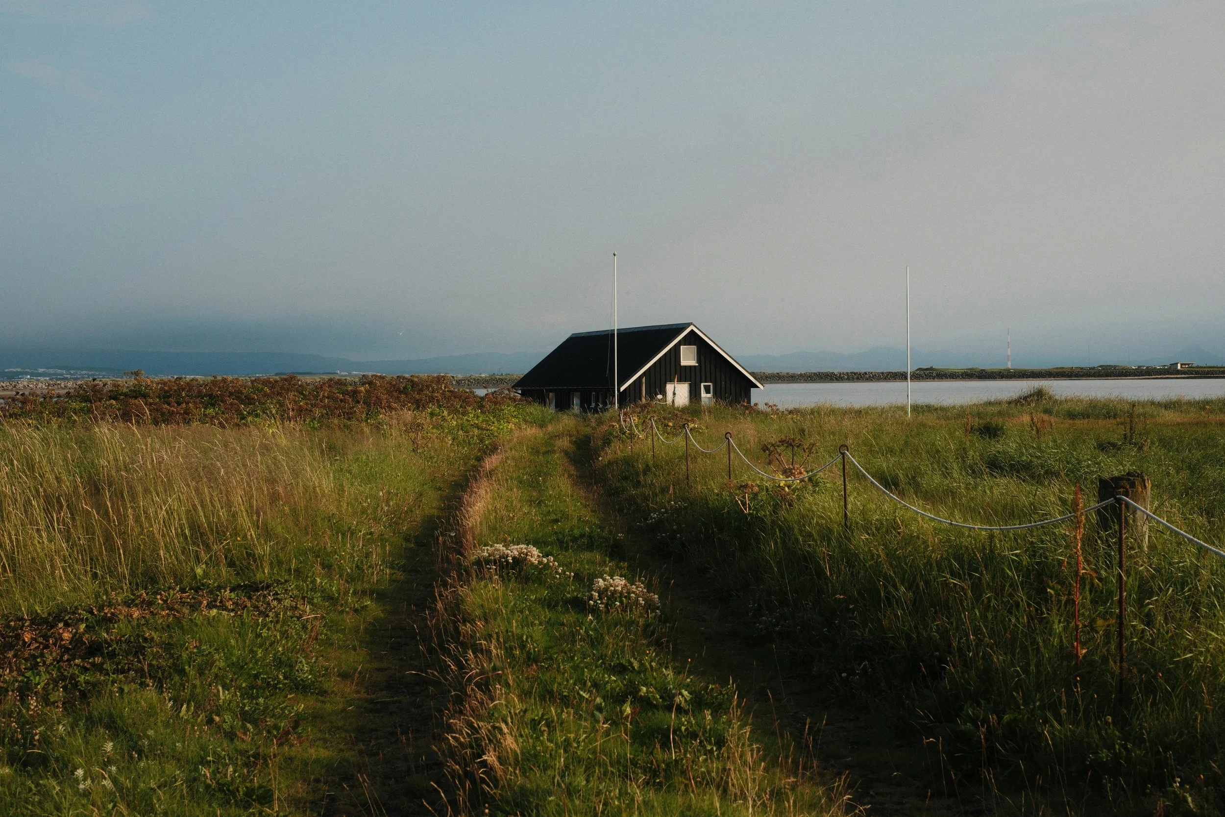 Black wooden cottage beside a coastal lagoon, surrounded by tall grass with mountains under a cloudy sky