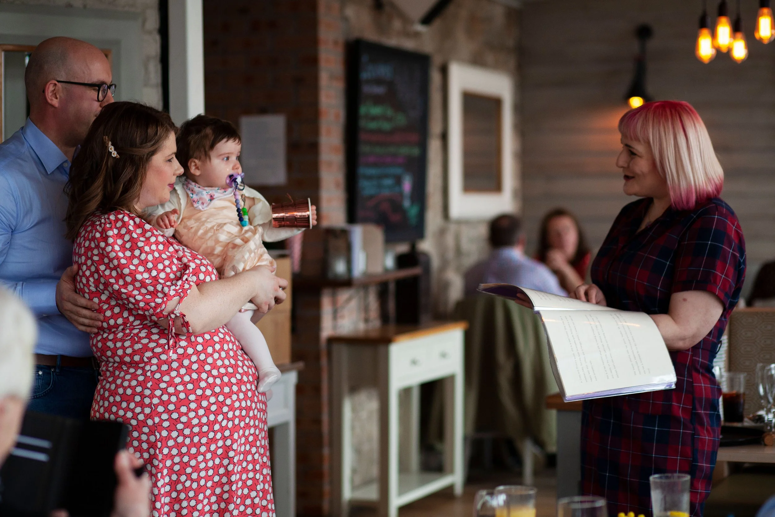 Sarah is conducting a naming ceremony. 2 parents are holding their daughter, Sarah is facing them and reading to them, the baby and Sarah are looking at each other and smiling.