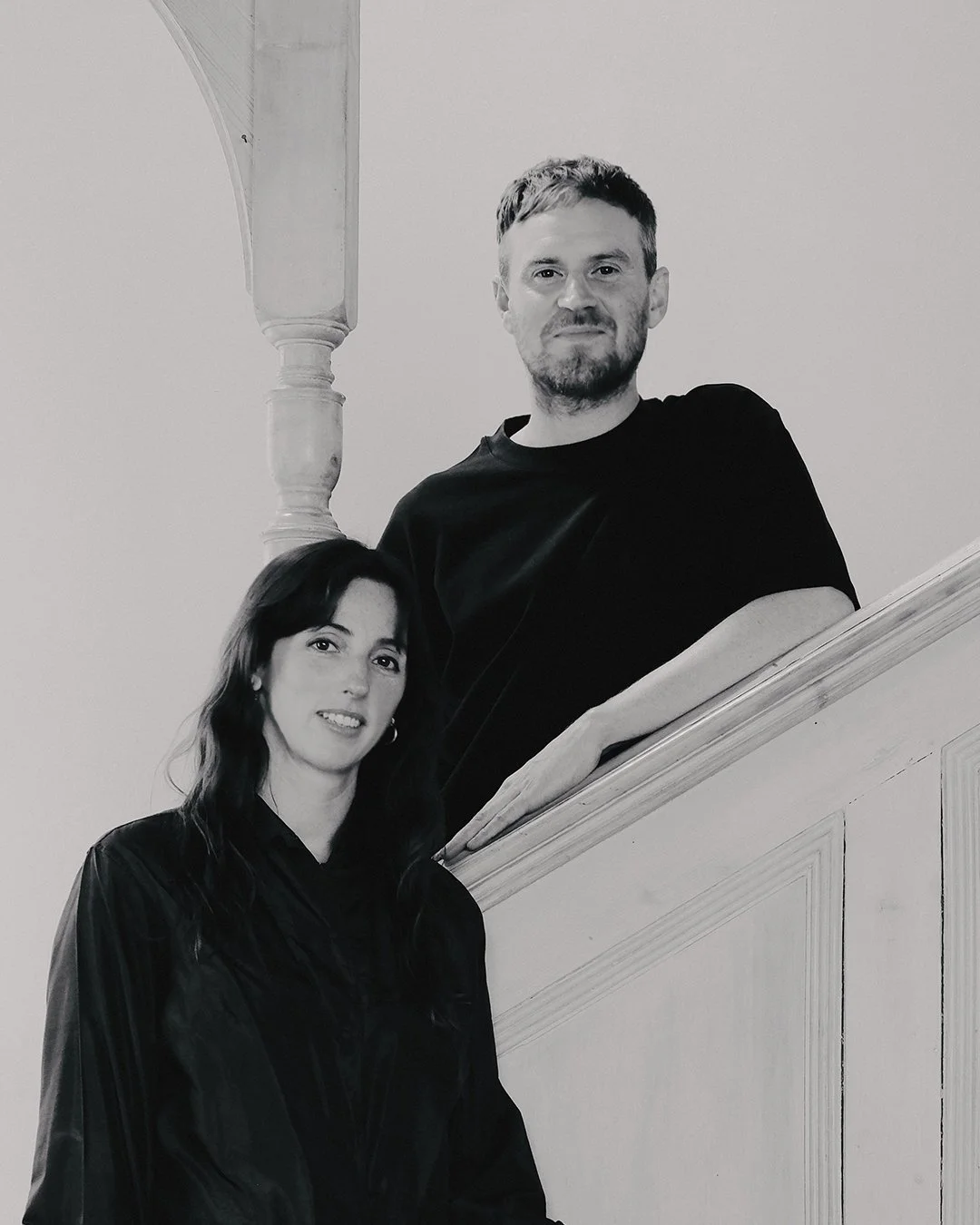 A black and white photo of a man and a woman posing on a staircase; the man is standing above and the woman is sitting on a lower step. The man has short hair, a beard, and is wearing a black t-shirt. The woman has long dark hair, is smiling slightly, and is wearing a dark jacket.