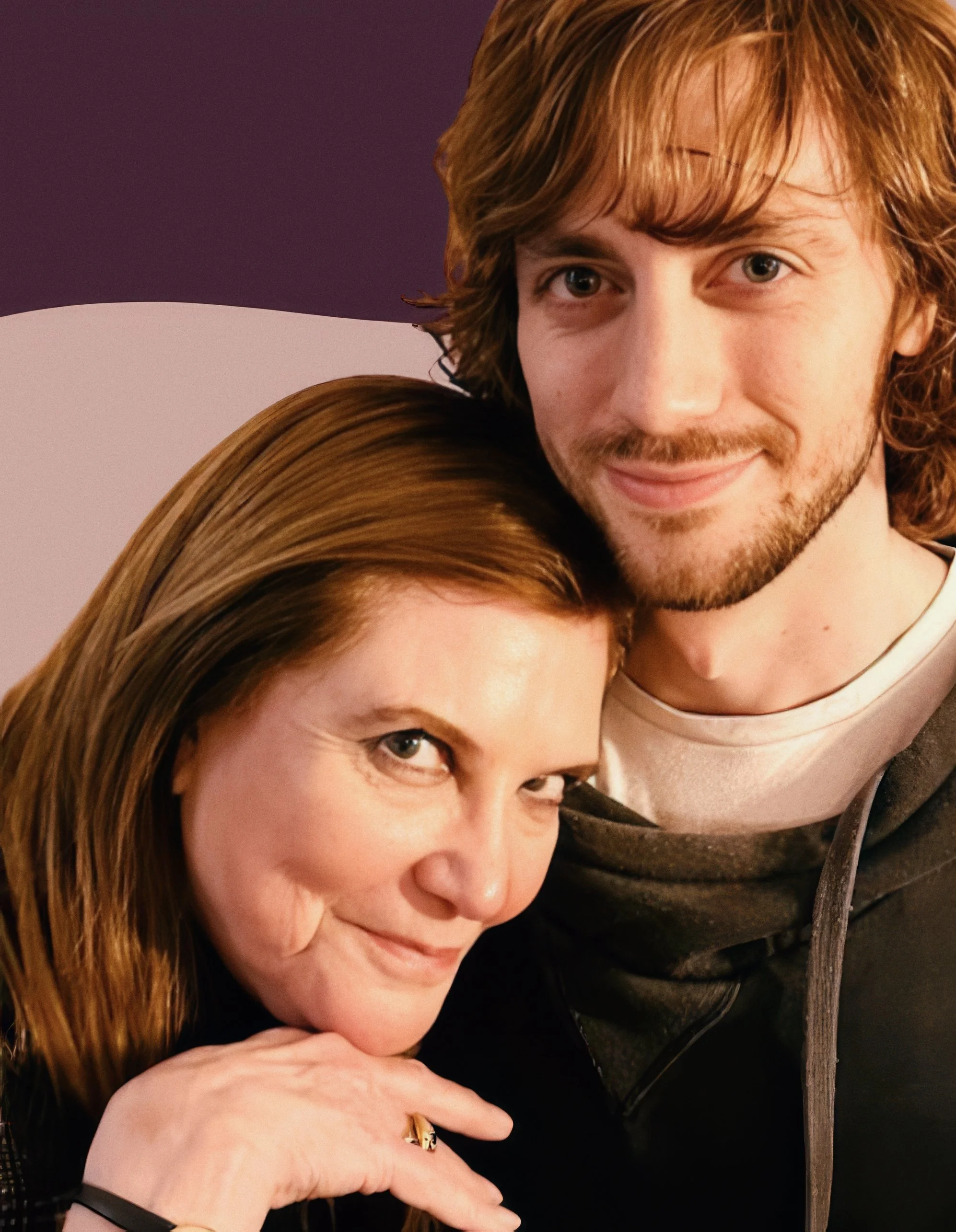 Close-up portrait of a smiling woman with long reddish-brown hair and a young man with curly brown hair, both leaning close together, with a purple background.