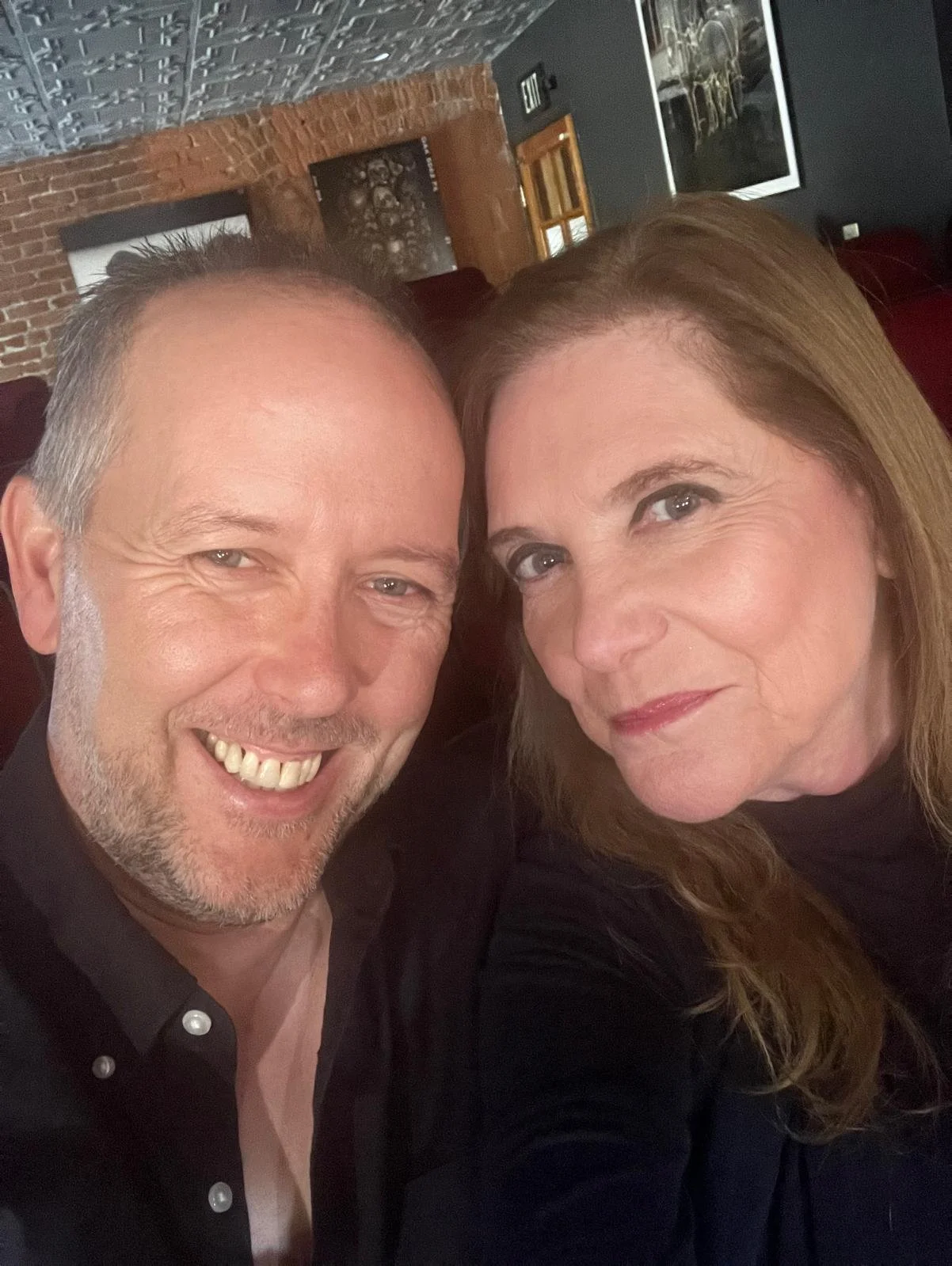 A smiling man and a woman with red hair taking a selfie together in a dimly lit indoor setting with brick walls and artwork in the background.