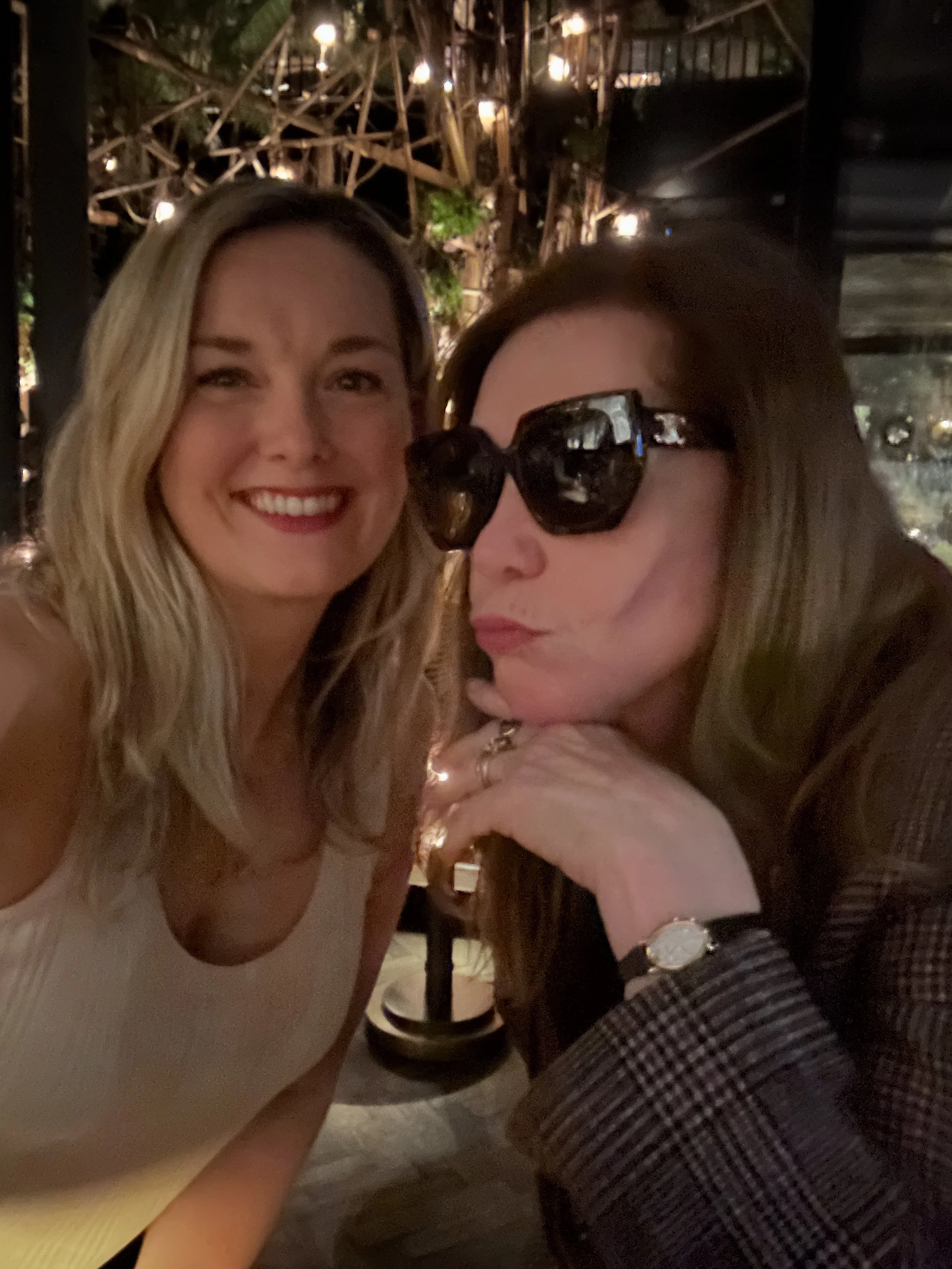 Two women taking a selfie at night, one smiling with blonde hair and the other wearing sunglasses with brown hair; background has warm lighting and decorative structures.