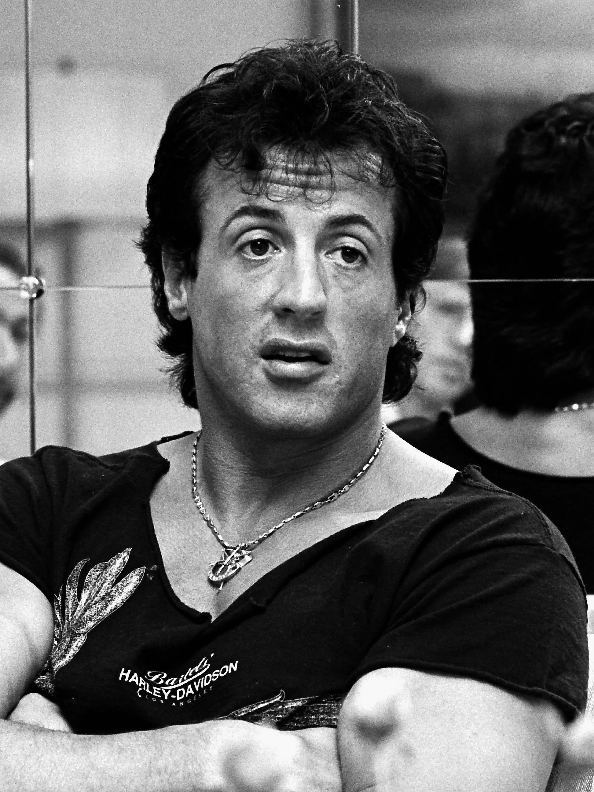Black and white photo of a man with dark, curly hair, wearing a Harley-Davidson t-shirt and a necklace, looking slightly to the side with an expression of surprise or confusion.