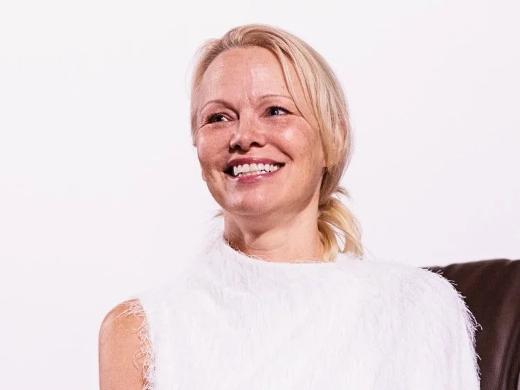 A woman with blonde hair smiling, wearing a white sleeveless top, sitting on a dark-colored chair against a plain white background.