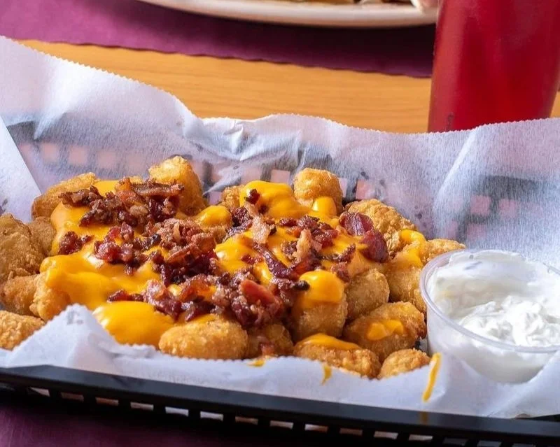 Fried chicken bites topped with melted cheese and bacon bits, served with a side of ranch dressing in a small cup on a black basket lined with parchment paper.