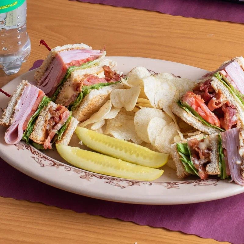 A plate with a club sandwich cut into quarters, showing layers of ham, turkey, lettuce, tomato, and bread. The plate also has potato chips and two pickle spears.