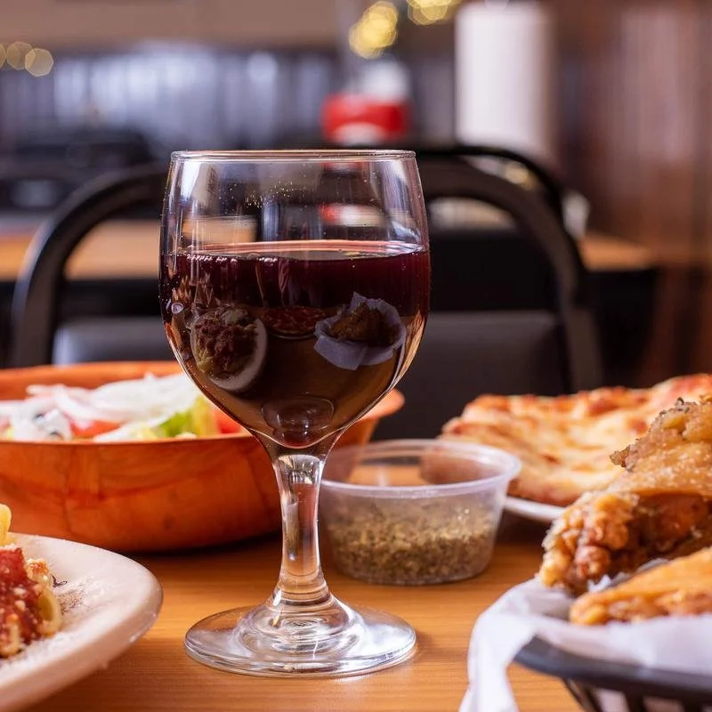 A glass of red wine on a table with various dishes including salad, fried chicken, and pizza in the background.