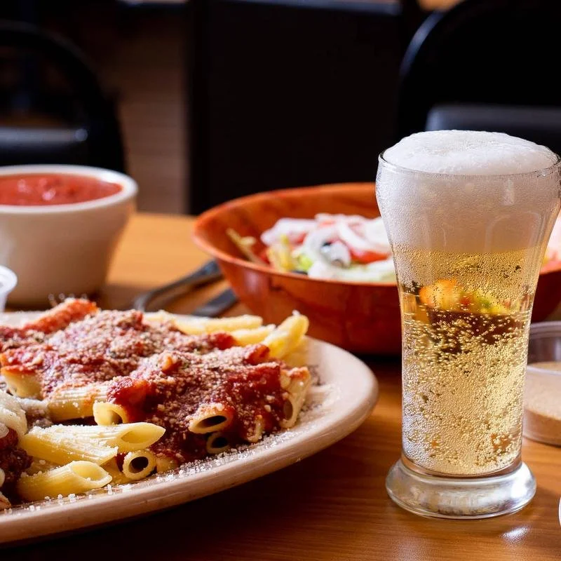 A plate of penne pasta with tomato sauce and grated cheese, a glass of sparkling beverage with a foamy top, a bowl of salad with dressing, and a bowl of tomato sauce on a dining table.