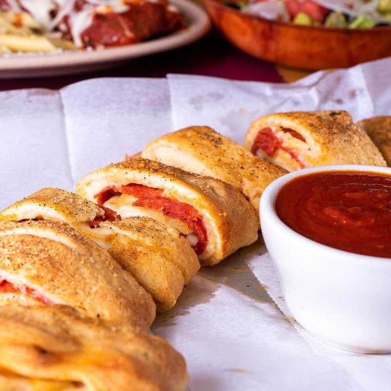 Four chicken cordon bleu rolls with breaded coating on white parchment, served with a cup of marinara sauce, on a table with other dishes in the background.
