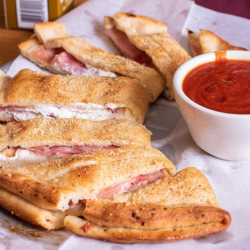 A plate of stuffed garbage bread sliced, with ham, cheese, and breading, served with a side of marinara sauce in a bowl.