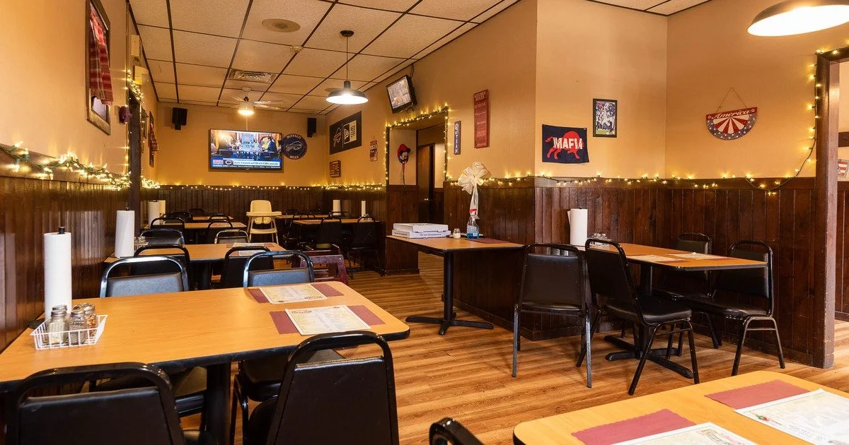 Empty restaurant or diner with wooden floors, tables set with menus, and dark chairs. Decor includes string lights, posters on the walls, a TV, and a high chair in the background.