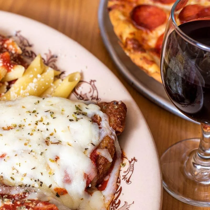 A plate with baked Italian dish, pasta with sauce, and breaded meat with cheese, accompanied by a glass of red wine and a slice of pepperoni pizza in the background.