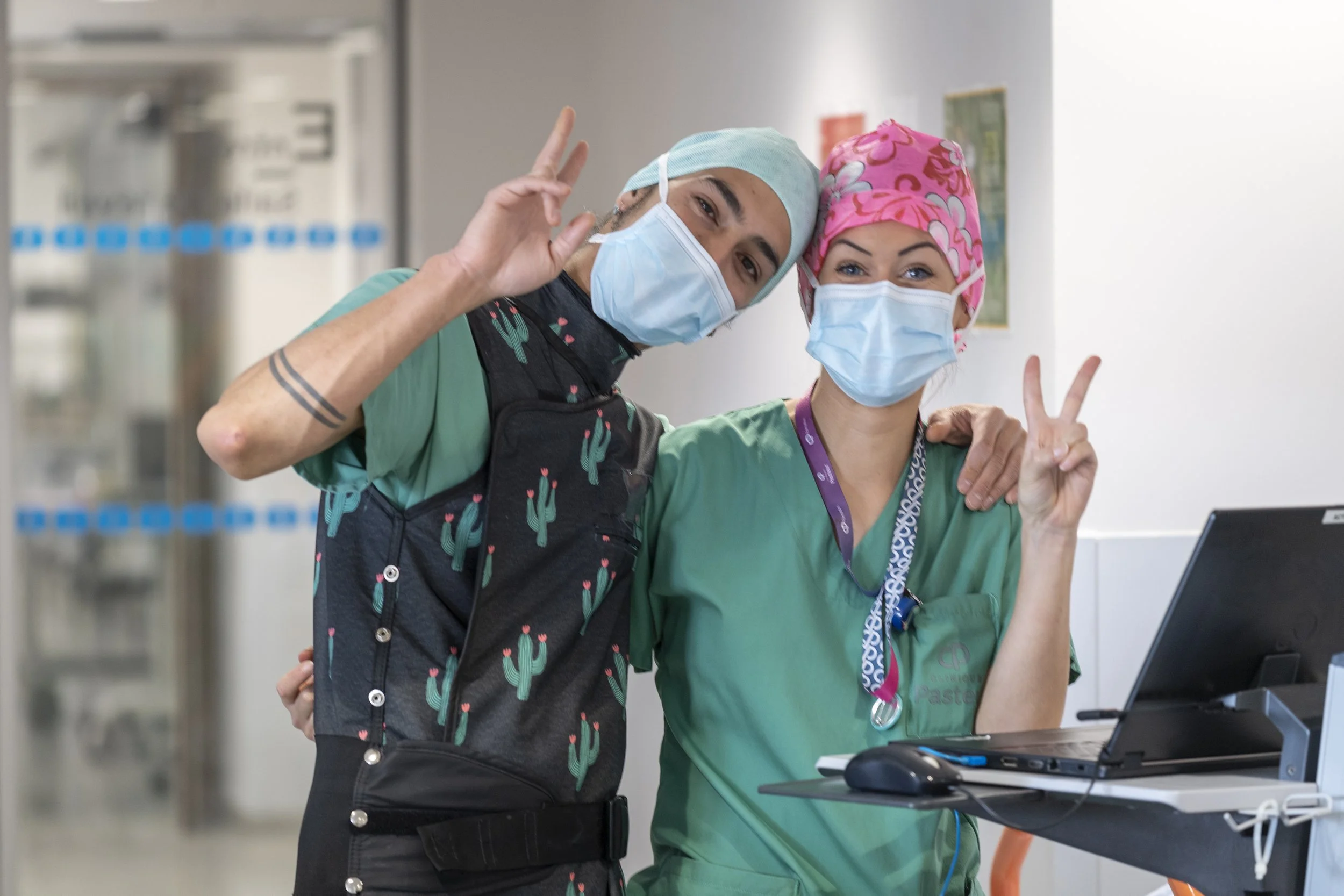 Deux professionnels de santé masqués en tenue chirurgicale, souriant et faisant le signe de la victoire dans un hôpital.