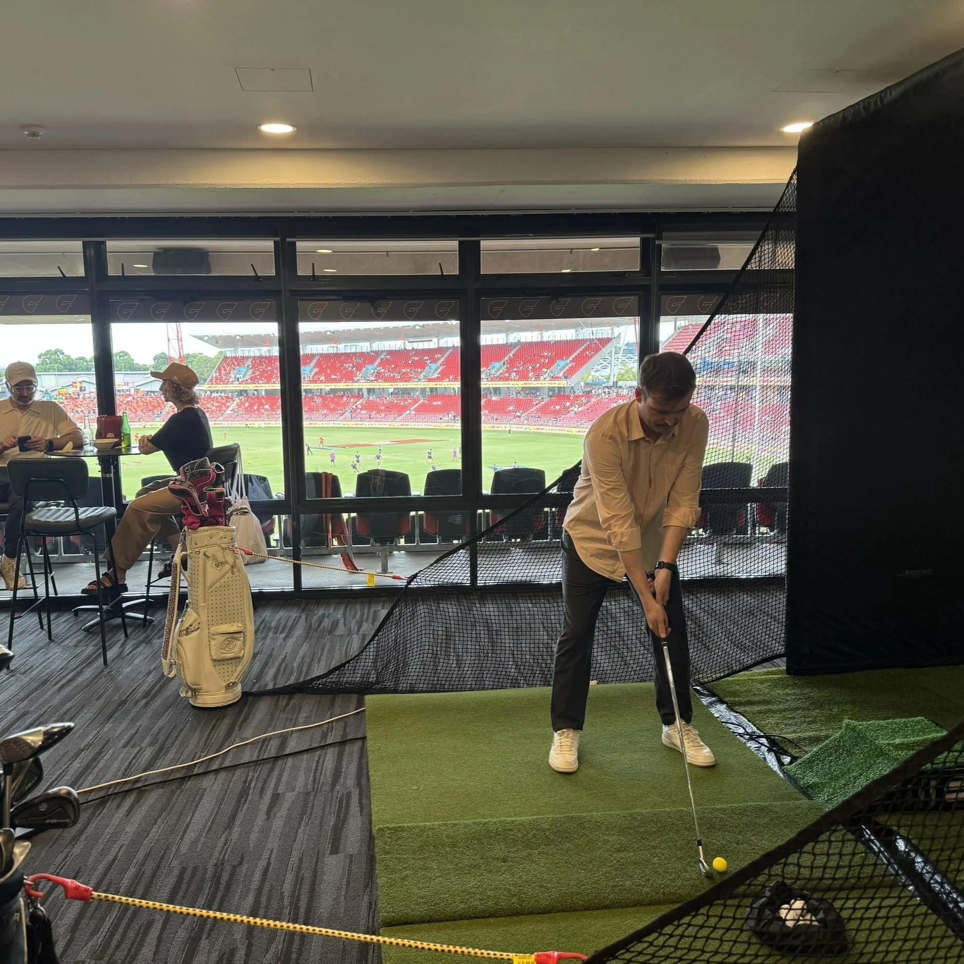 Participant using mobile golf simulator with AFL grandstand in the background of image.