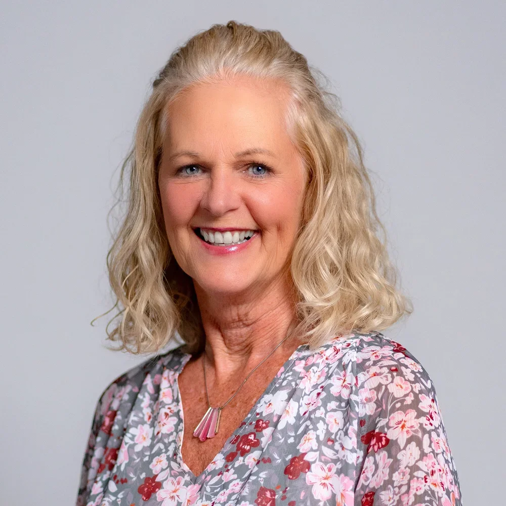 A smiling middle-aged woman with blonde curly hair, wearing a floral blouse and a pink necklace, against a plain light gray background.