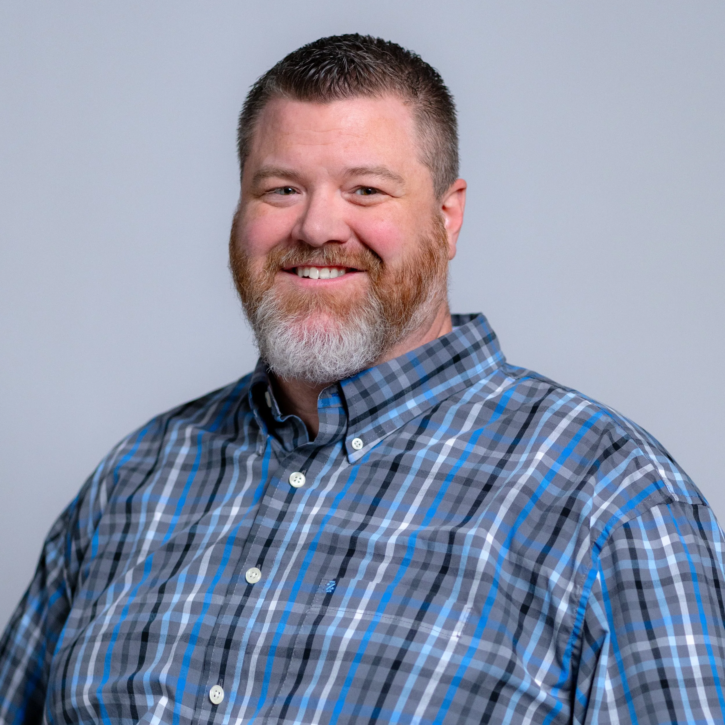 A man with a beard and short hair smiling, wearing a blue and gray plaid button-up shirt, against a plain light gray background.