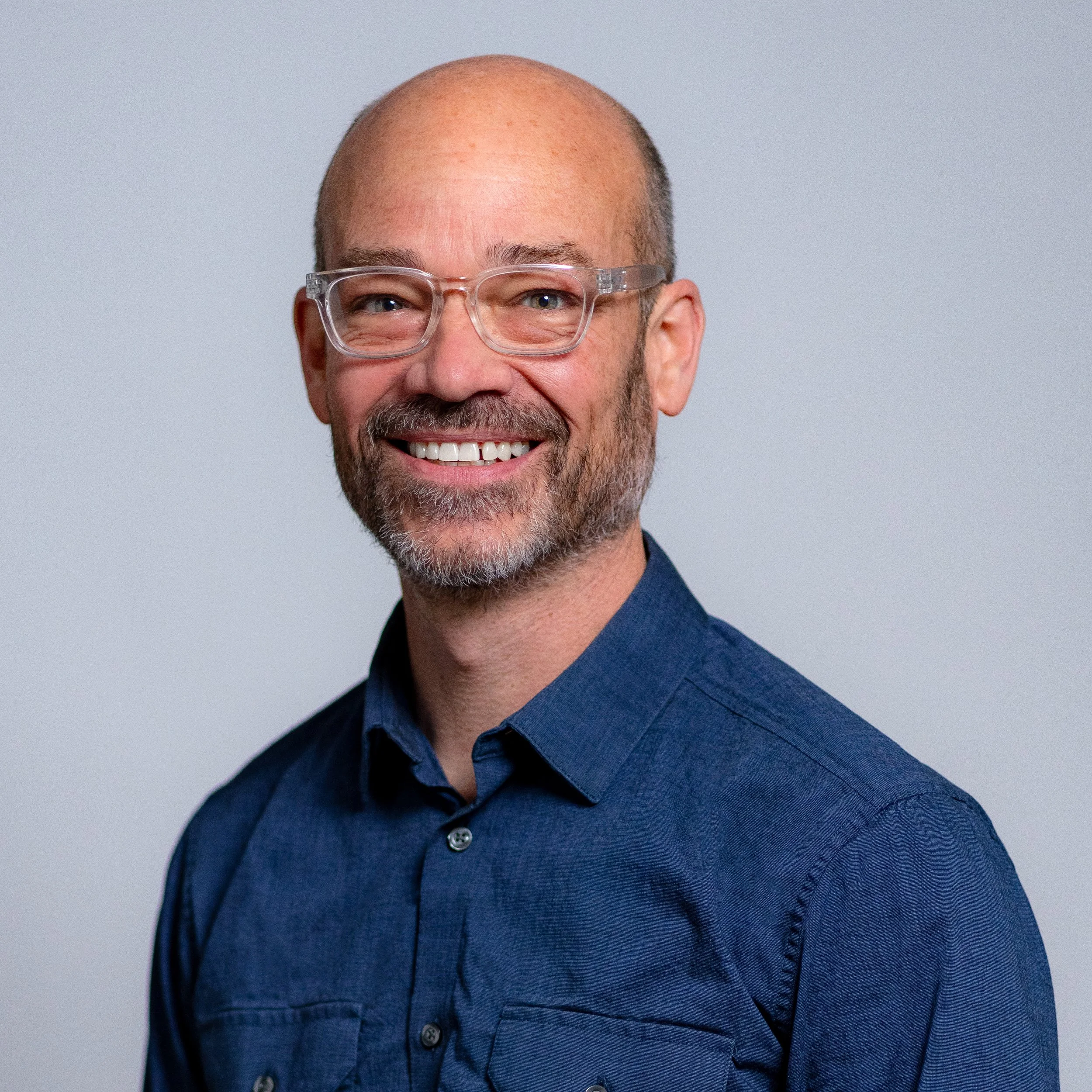 A smiling man with glasses, a beard, and a mustache, wearing a blue button-up shirt, against a plain light gray background.